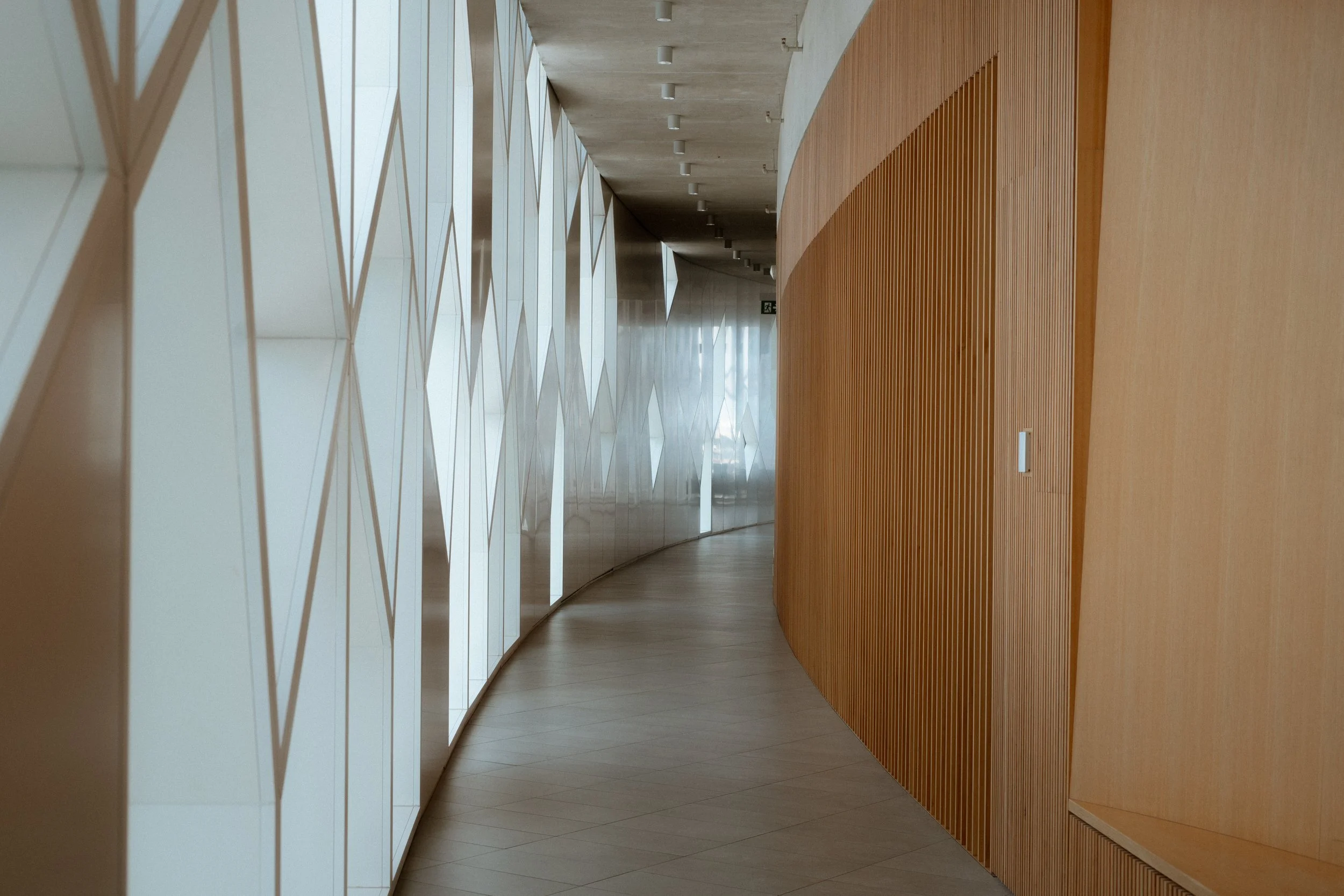Interior corridor of Calgary Central Library showing curved glass façade with geometric pattern alongside vertical wood wall enclosing the Great Reading Room.