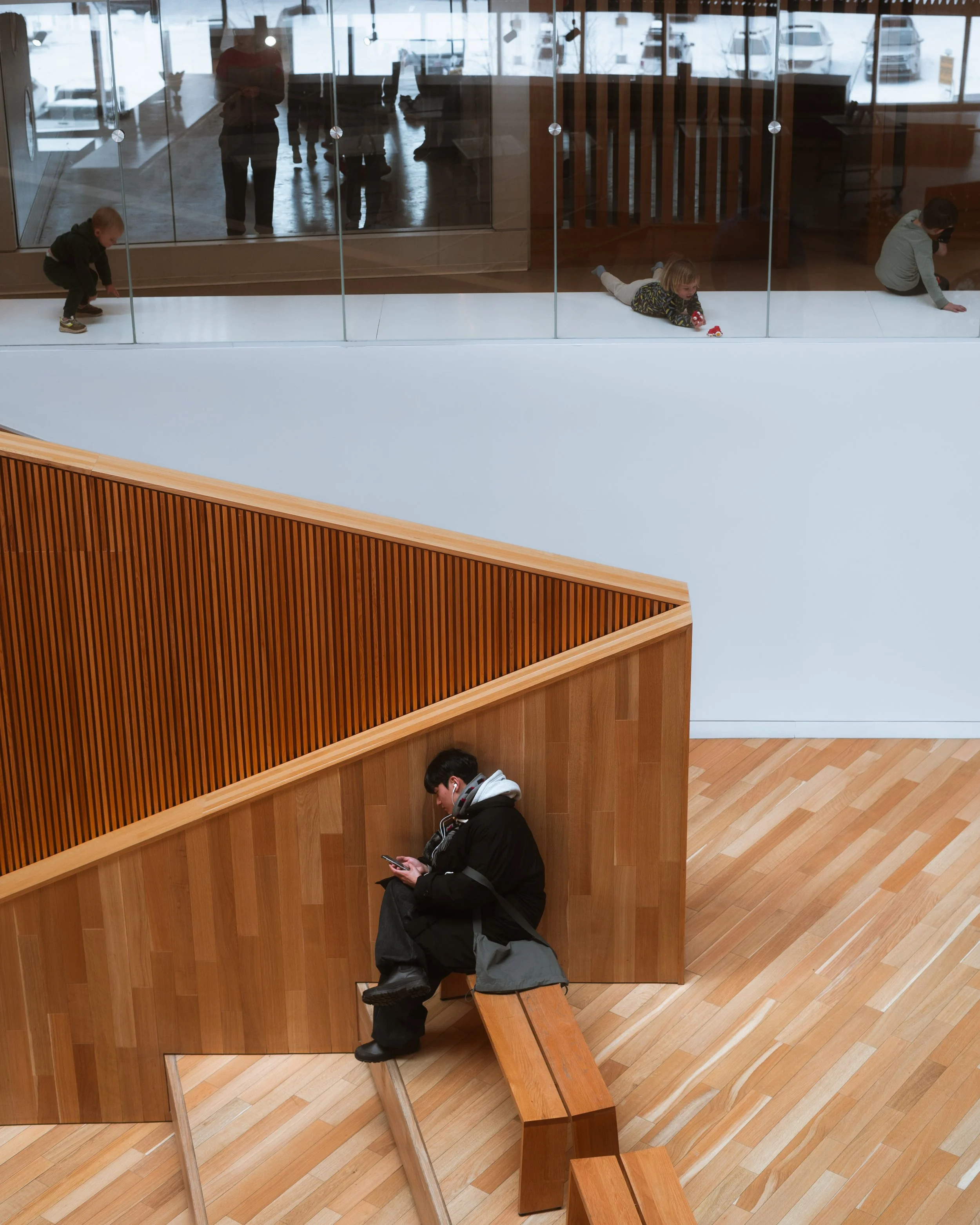 Calgary Central Library interior showing stepped wood seating in use at the entrance, with glass separating the children’s area above from the main circulation space.