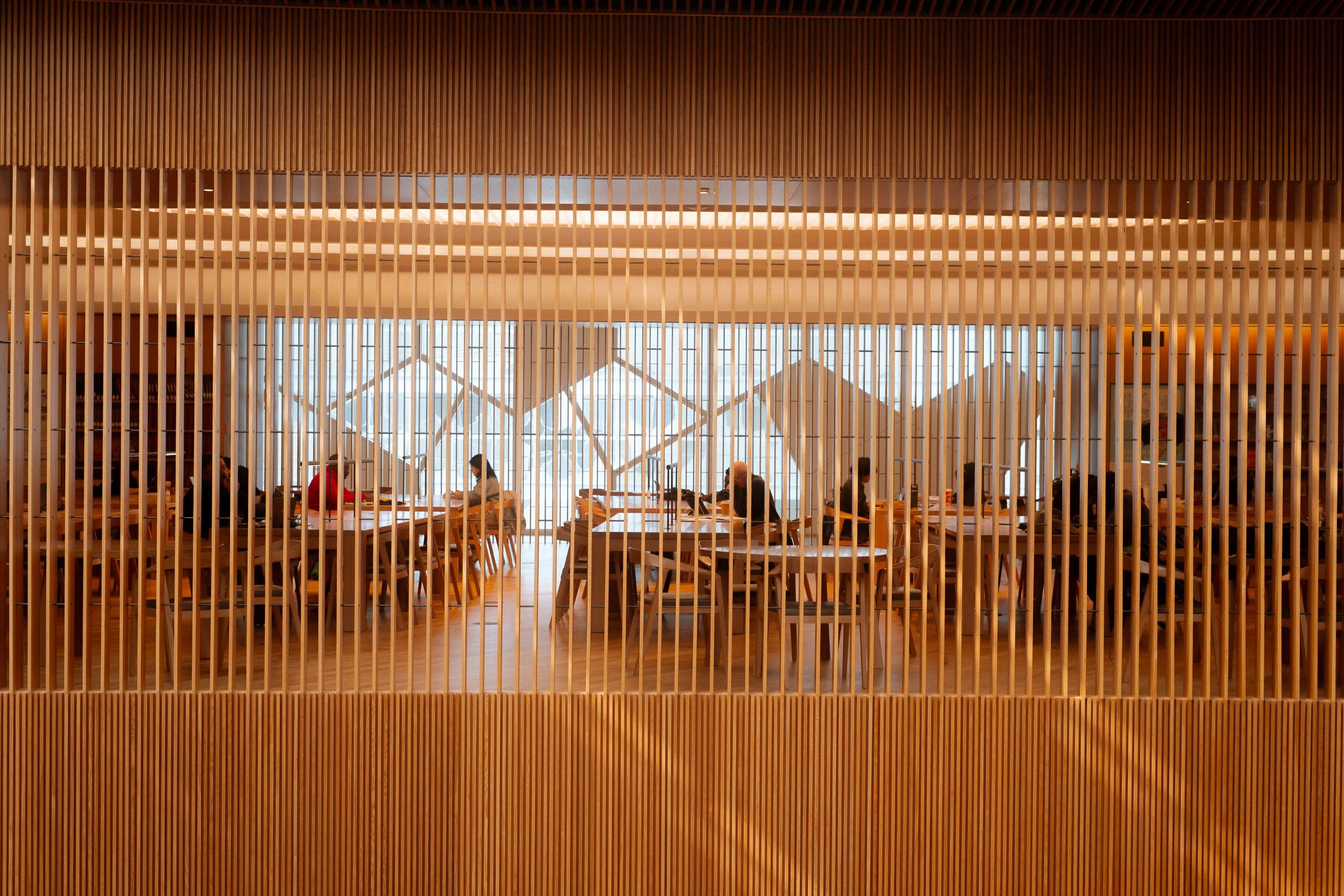 Calgary Central Library Great Reading Room seen through vertical wood slats, showing study tables, filtered light, and layered separation between circulation and quiet space.