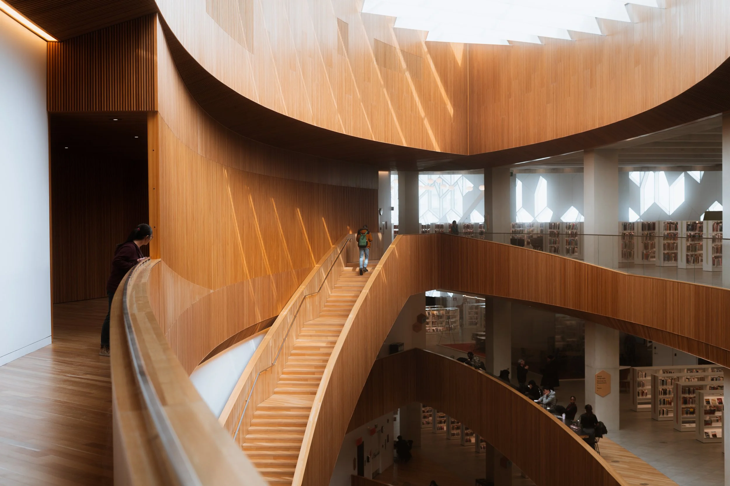 Calgary Central Library interior staircase with curved wood architecture and filtered daylight, connecting upper levels with the Great Reading Room entrance visible to the left.
