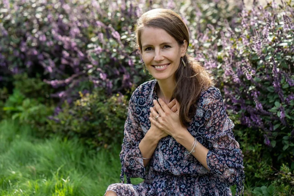 A woman with long brown hair smiling and holding her hands over her heart, sitting on grass in front of a bush with purple flowers.