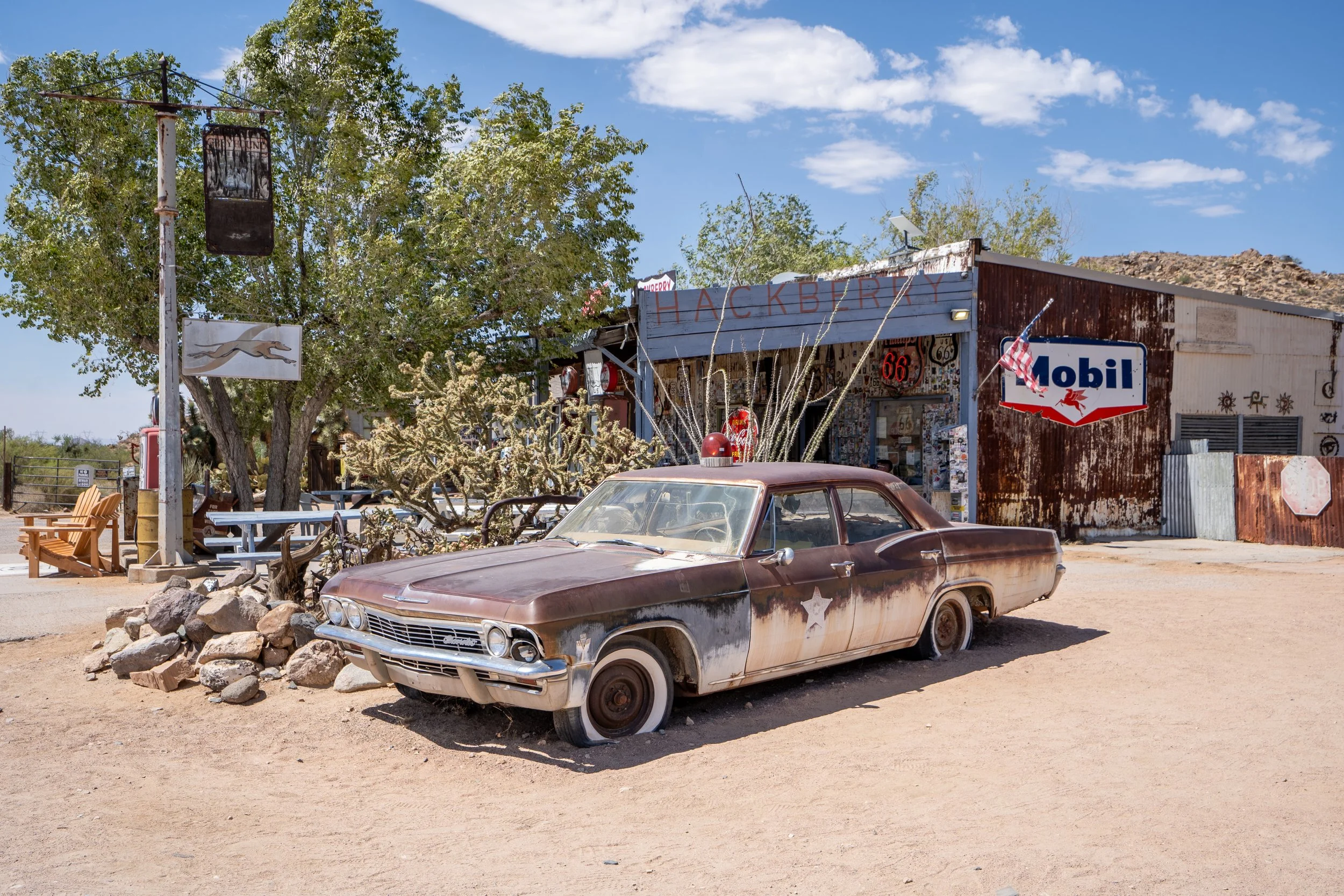 A vintage car parked in front of a small roadside shop with signs that read 'Hackberry' and 'Mobil'. There is a large tree, a rock pile, and a blue sky with scattered clouds in the background.