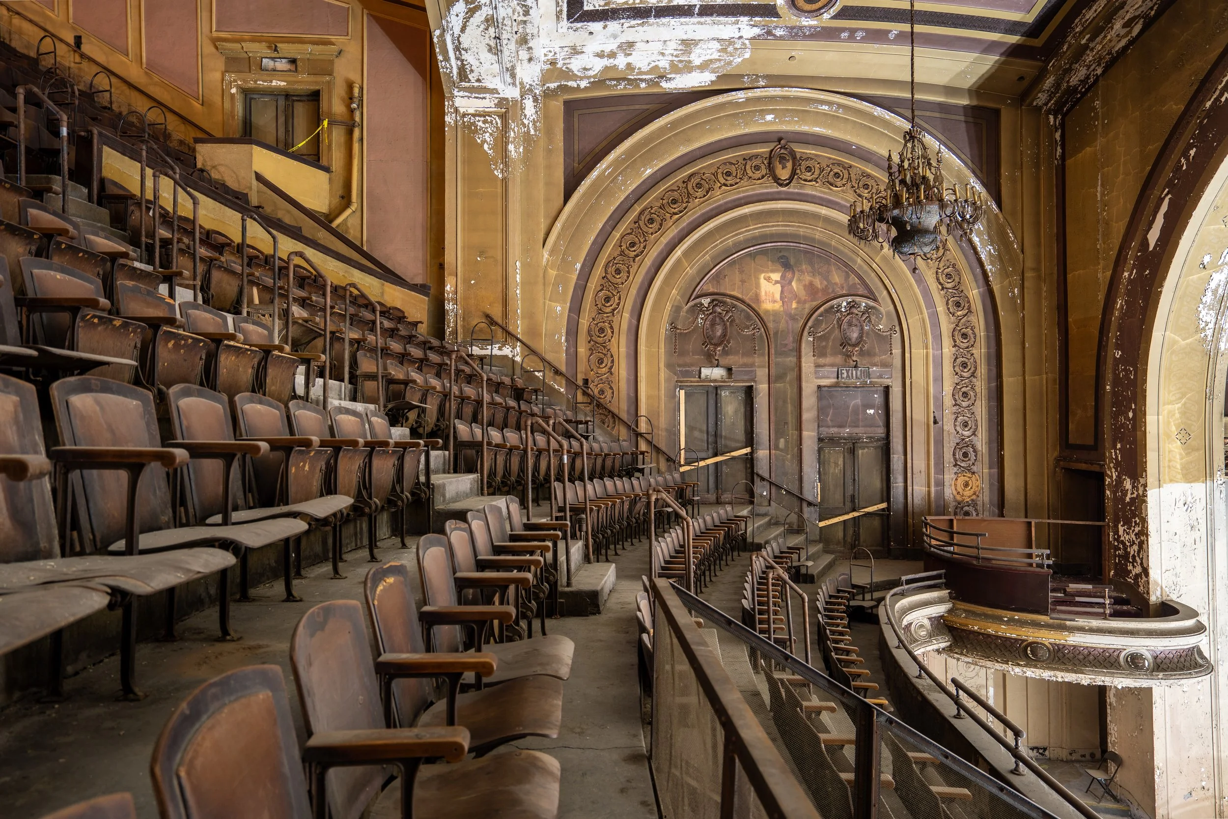 An abandoned theater with peeling paint on walls, dusty brown seats, ornate archways, and a chandelier hanging from the ceiling.