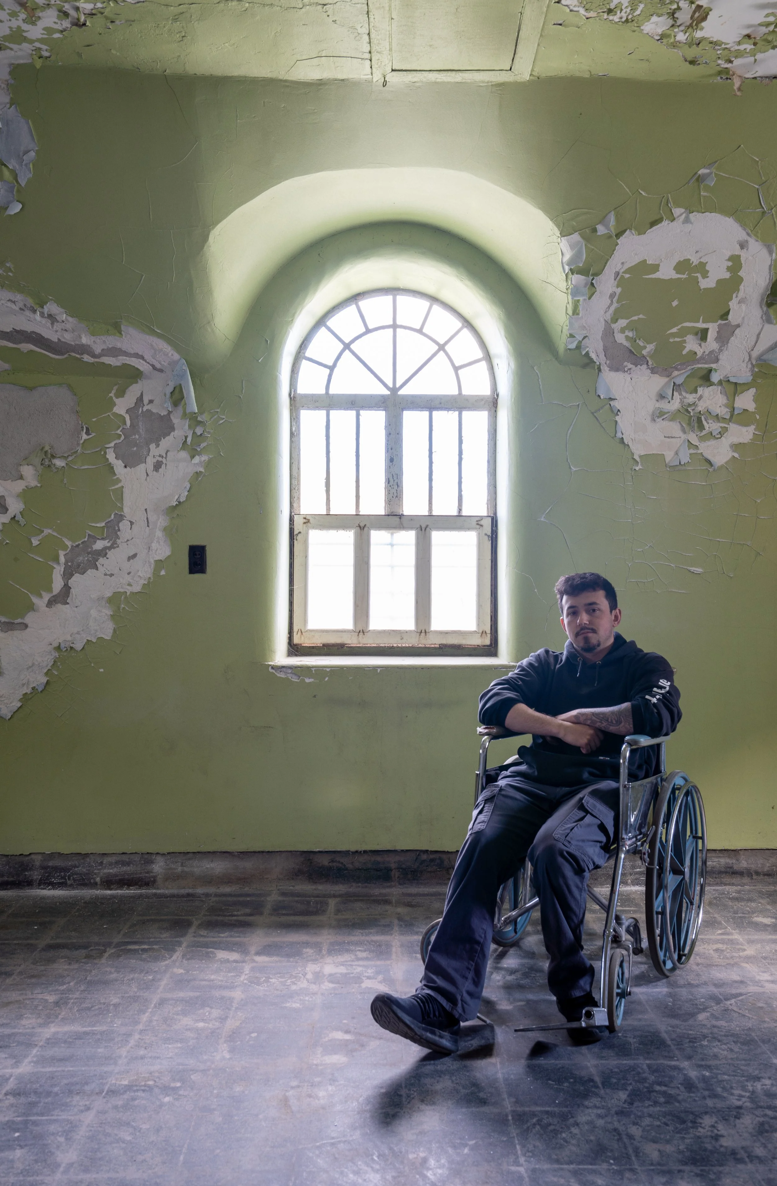 Young man in a wheelchair with crossed arms sitting in an abandoned, dilapidated room with peeling paint and an arched window.