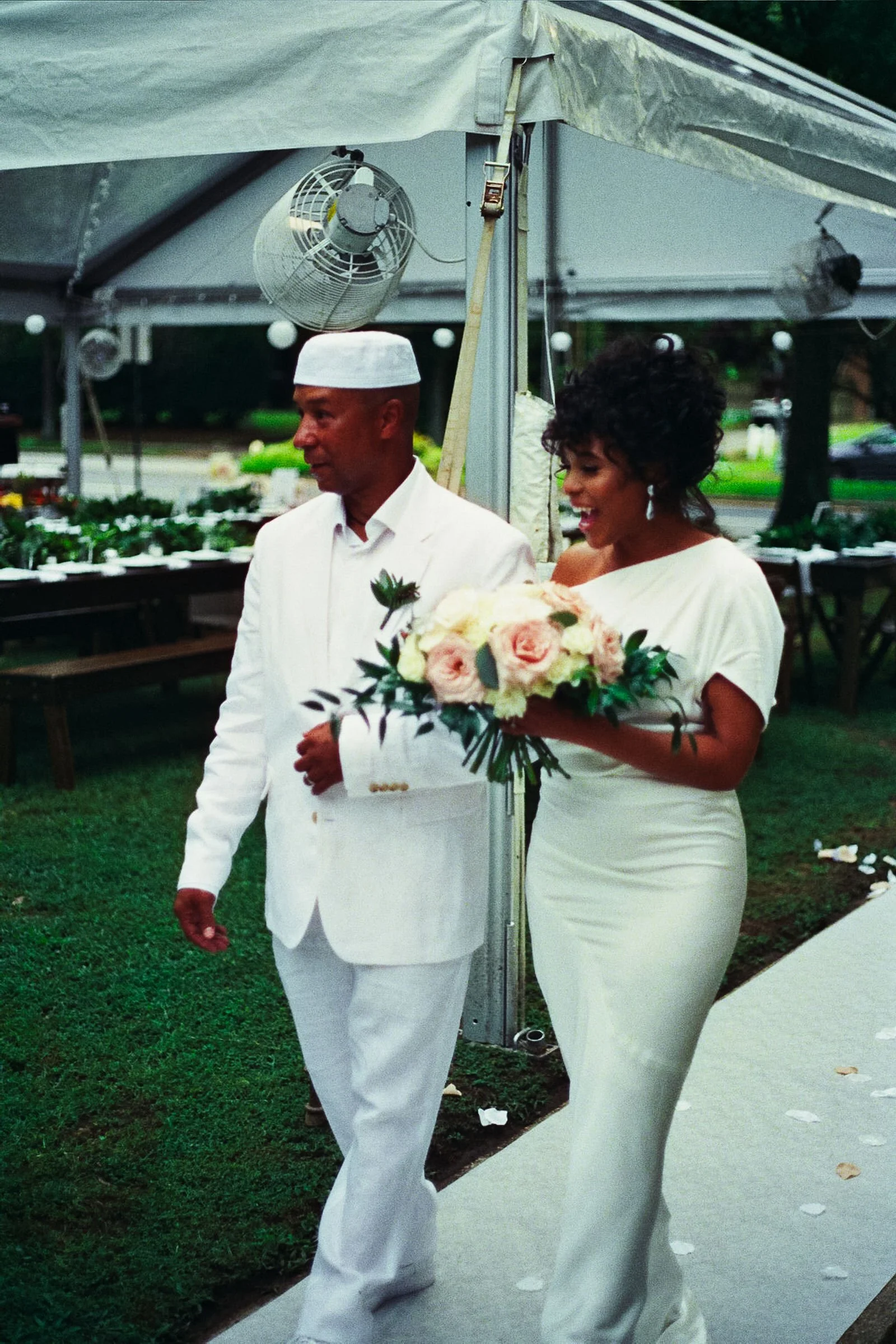 A woman in a white wedding dress holding a bouquet of flowers walking with a man in a white suit under a tent at an outdoor wedding reception.