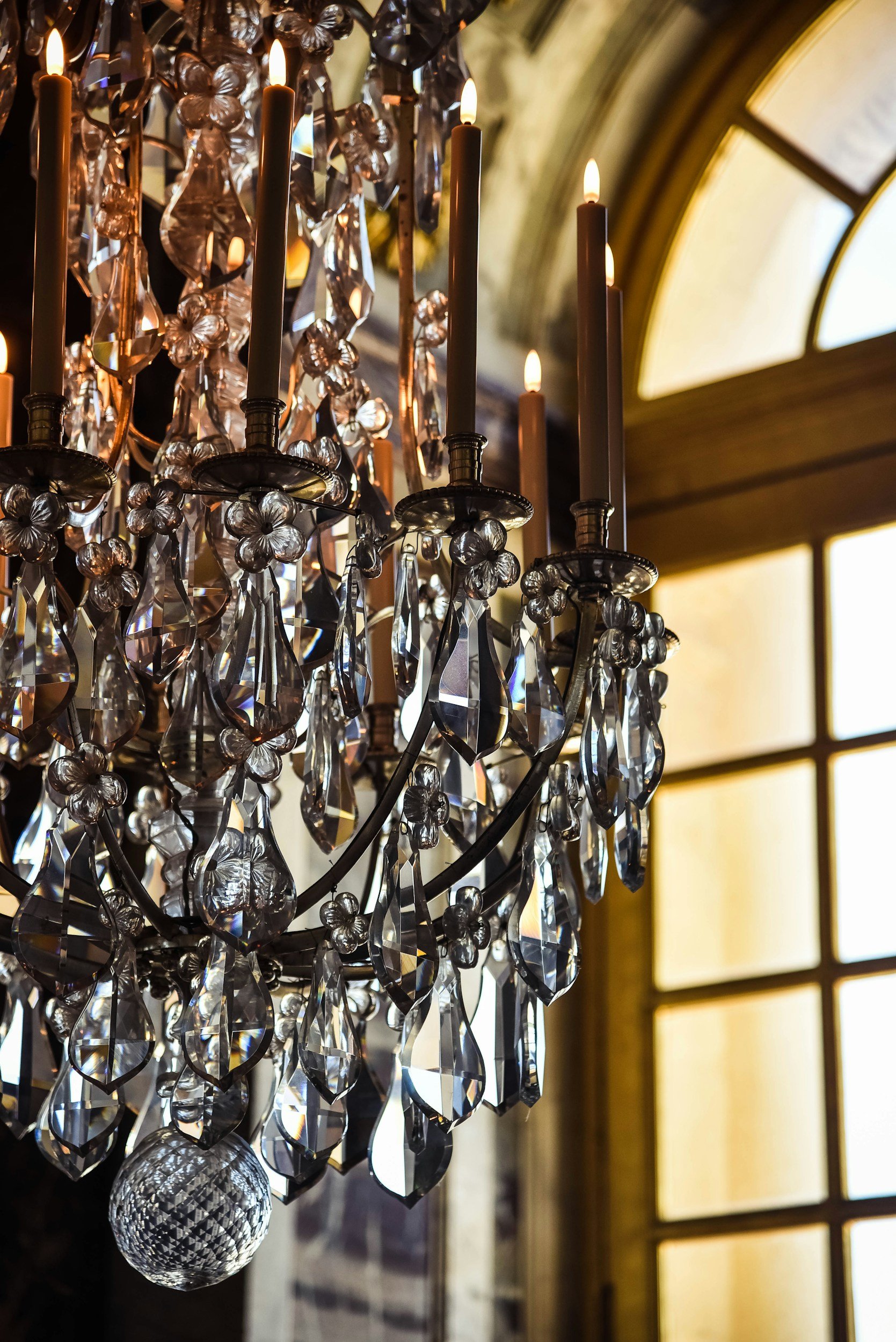 Close-up of a crystal chandelier with lit candles, hanging near a large arched window with a yellowish background.