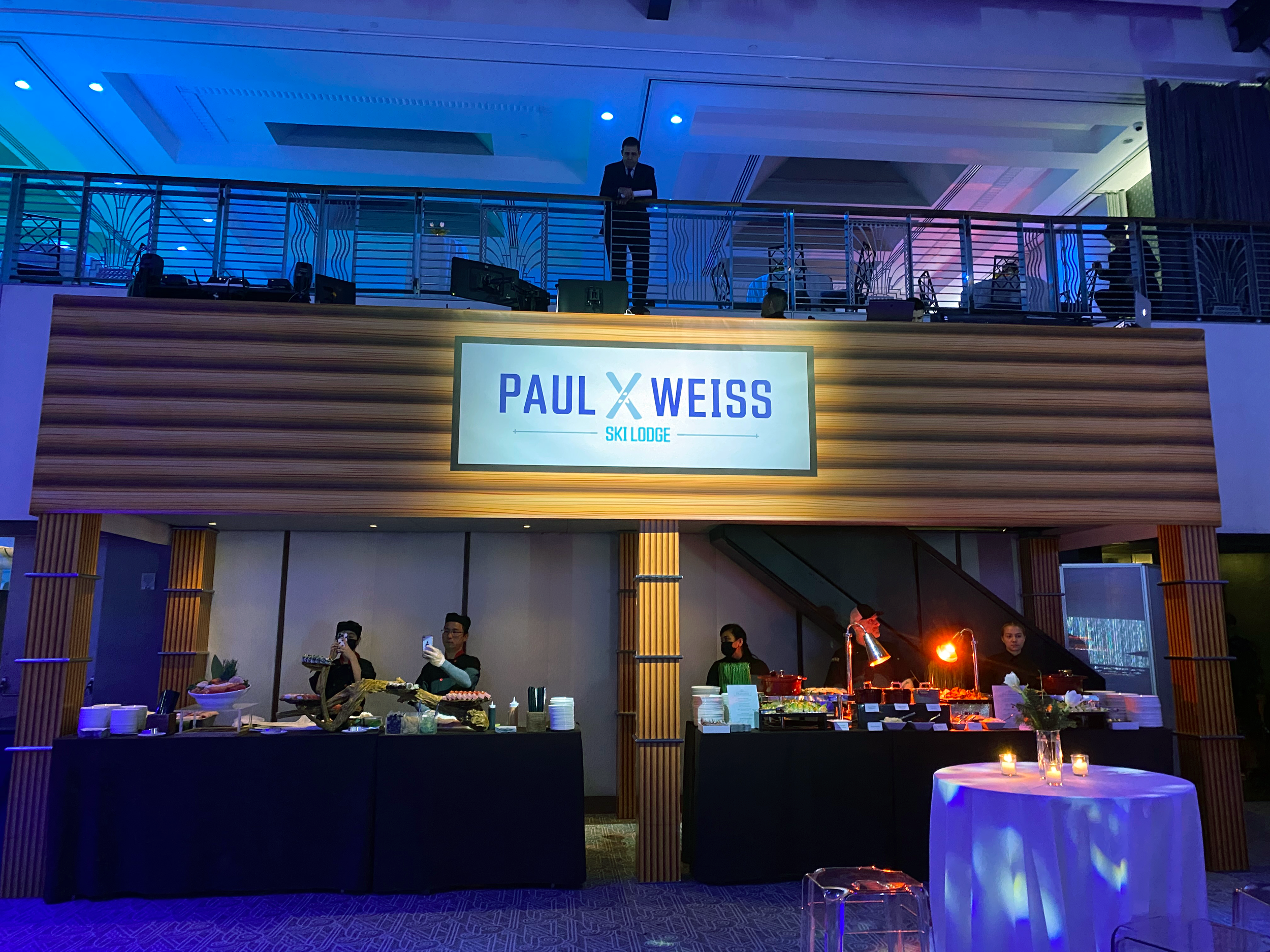 Event space with a large digital sign that reads 'PAUL WEISS SKI LODGE,' with a buffet table below, people serving food and taking photos, and a second level balcony with a person standing behind a railing.