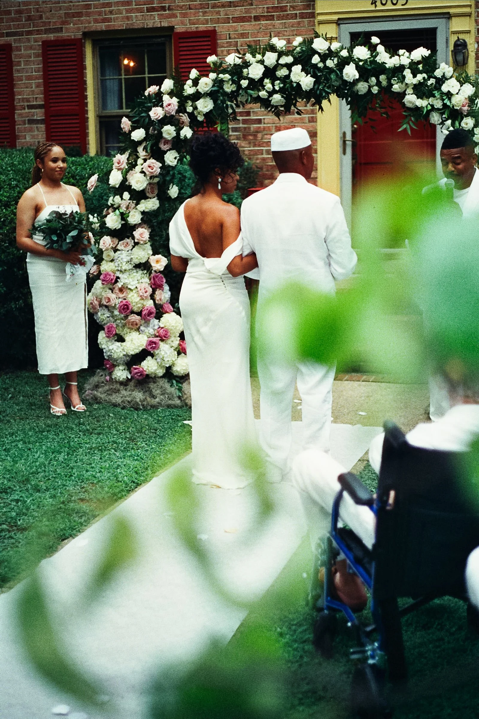 A couple getting married outdoors in front of a floral arch, with a woman holding a bouquet and a man in light-colored clothing, surrounded by greenery and a brick house.