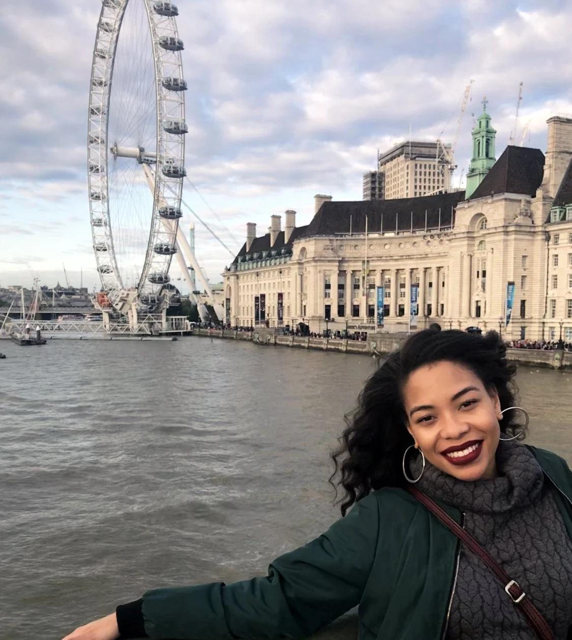 A smiling woman with dark curly hair, wearing hoop earrings, a dark green jacket, and a gray turtleneck sweater, poses near the riverfront with the London Eye and historic building behind her in London, England.