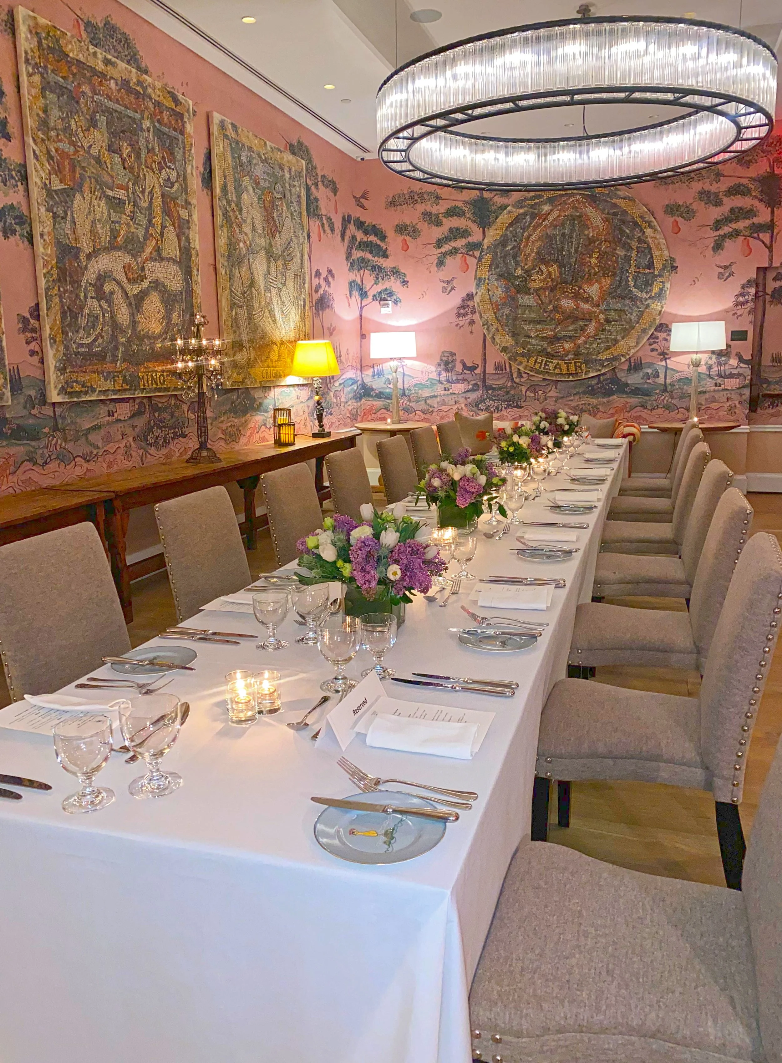 Elegant dining room set for a formal event with a long white tablecloth, floral centerpieces, and place settings, in a room with pink walls, large tapestry art, and a modern chandelier.