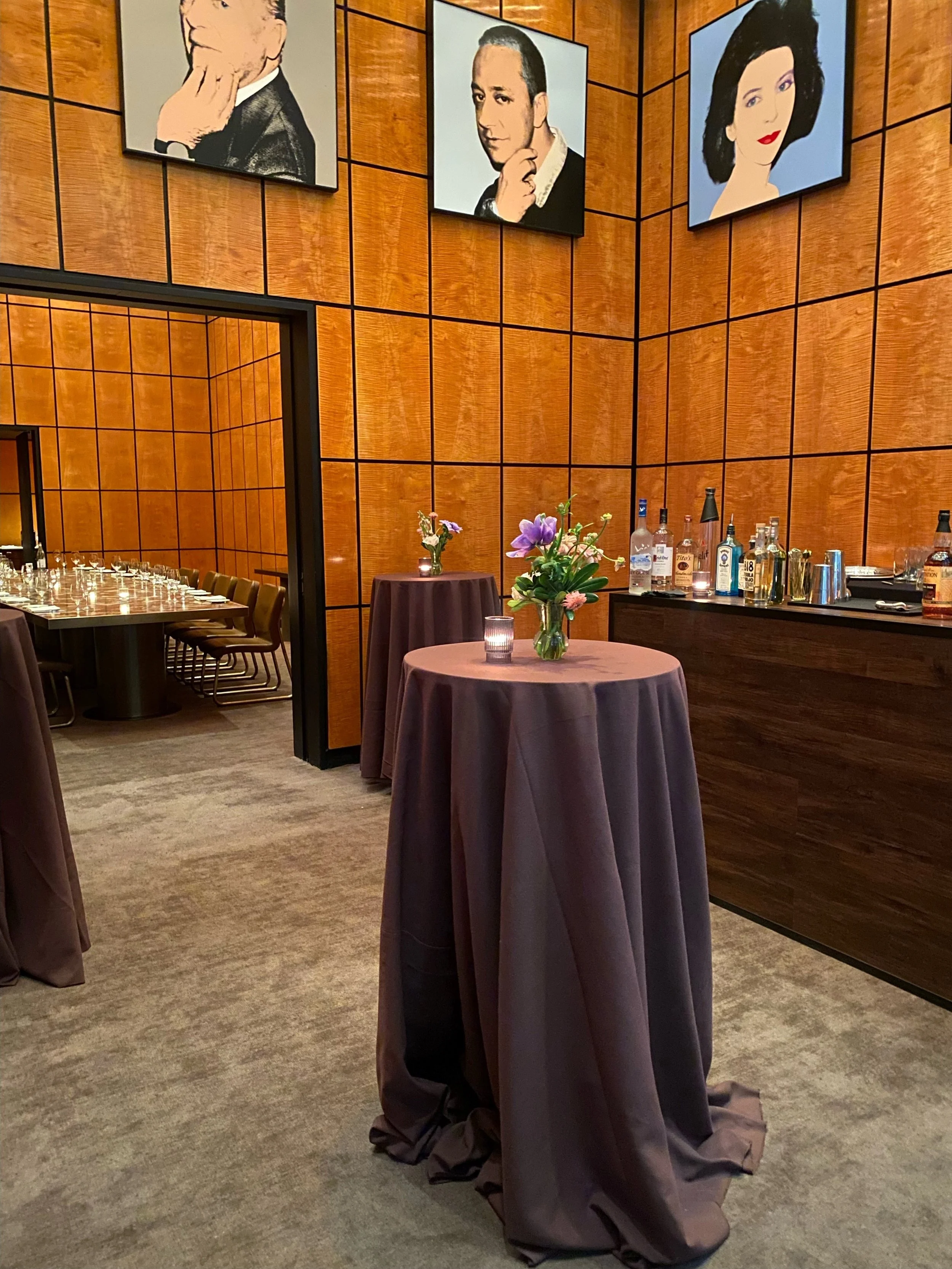 A bar area with brown covered high-top tables, a display of liquor bottles, and portraits of people on the wooden wall.