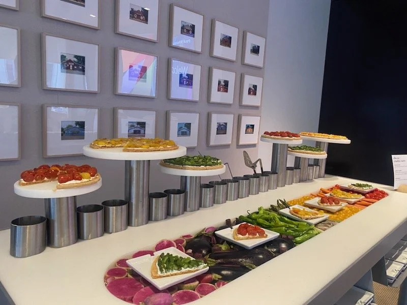 Display of sliced vegetables and toppings on a table, with small pizza slices on elevated platforms, set against a wall of framed photographs.