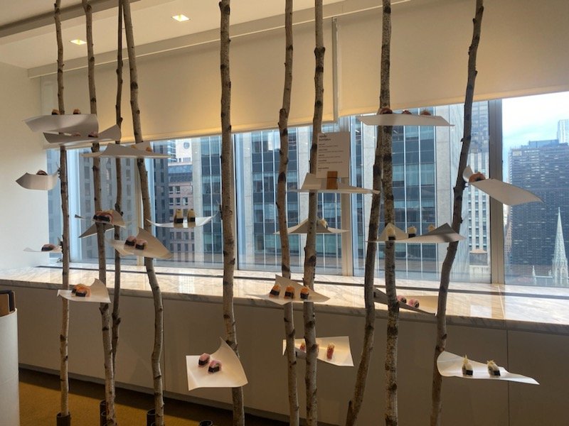 An indoor art display with vertical wooden sticks used as a partition, with paper boats and small objects placed on top of papers attached to the sticks. Cityscape view of tall buildings visible through large windows in the background.