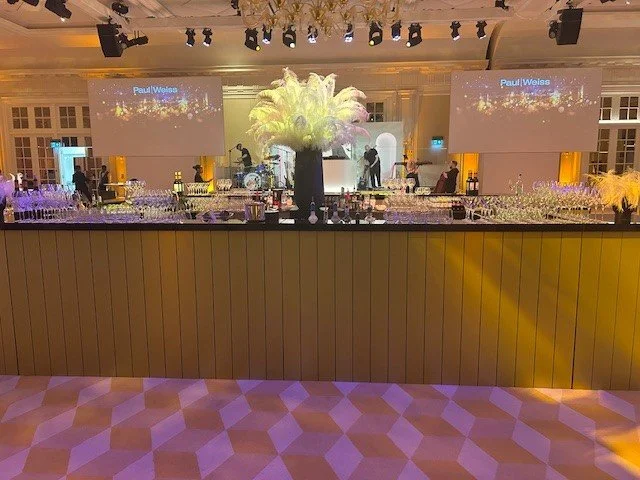 Event hall with a bar setup, large feathered centerpiece, and screens displaying 'Paul Weiss.'