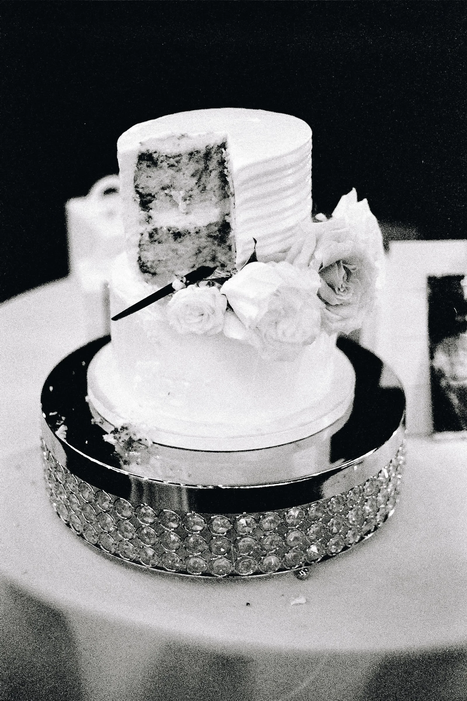 A black and white photograph of a multi-tiered wedding cake decorated with roses, a slice cut to reveal layers, and a cake topper, placed on a surface with a rhinestone trim around the base.