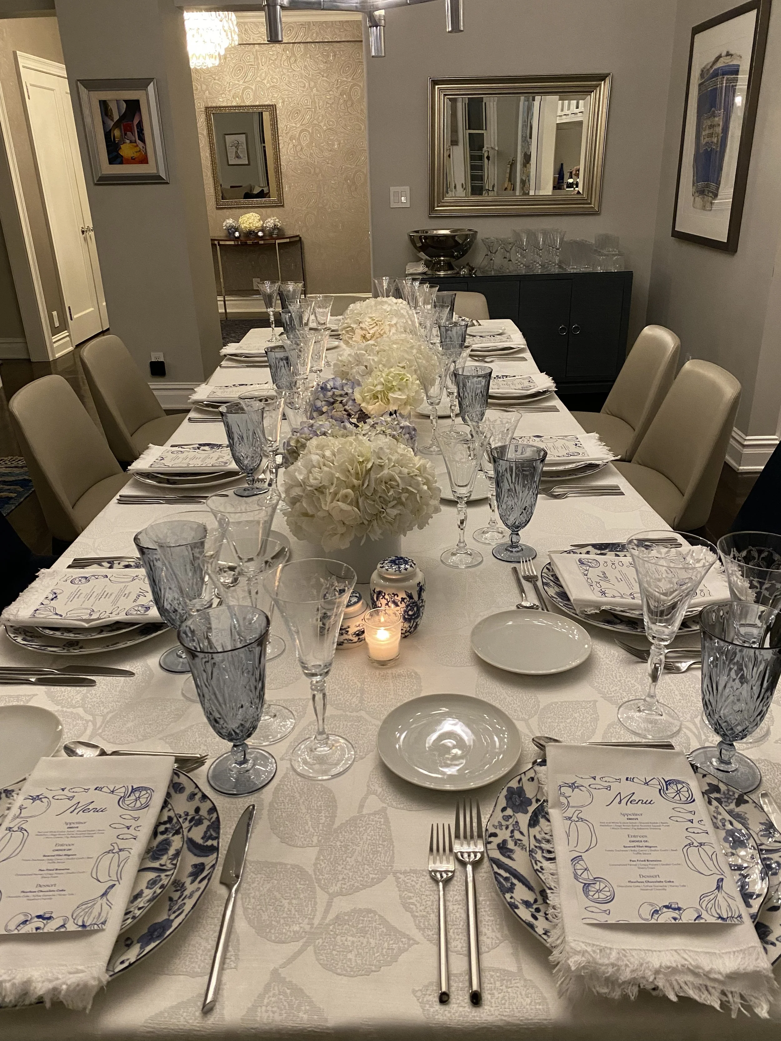 Elegant dining table set for a meal with white floral centerpieces, blue and white patterned plates, clear and blue glassware, silverware, and menu cards on a white tablecloth, in a well-decorated dining room with framed artwork and mirrors.