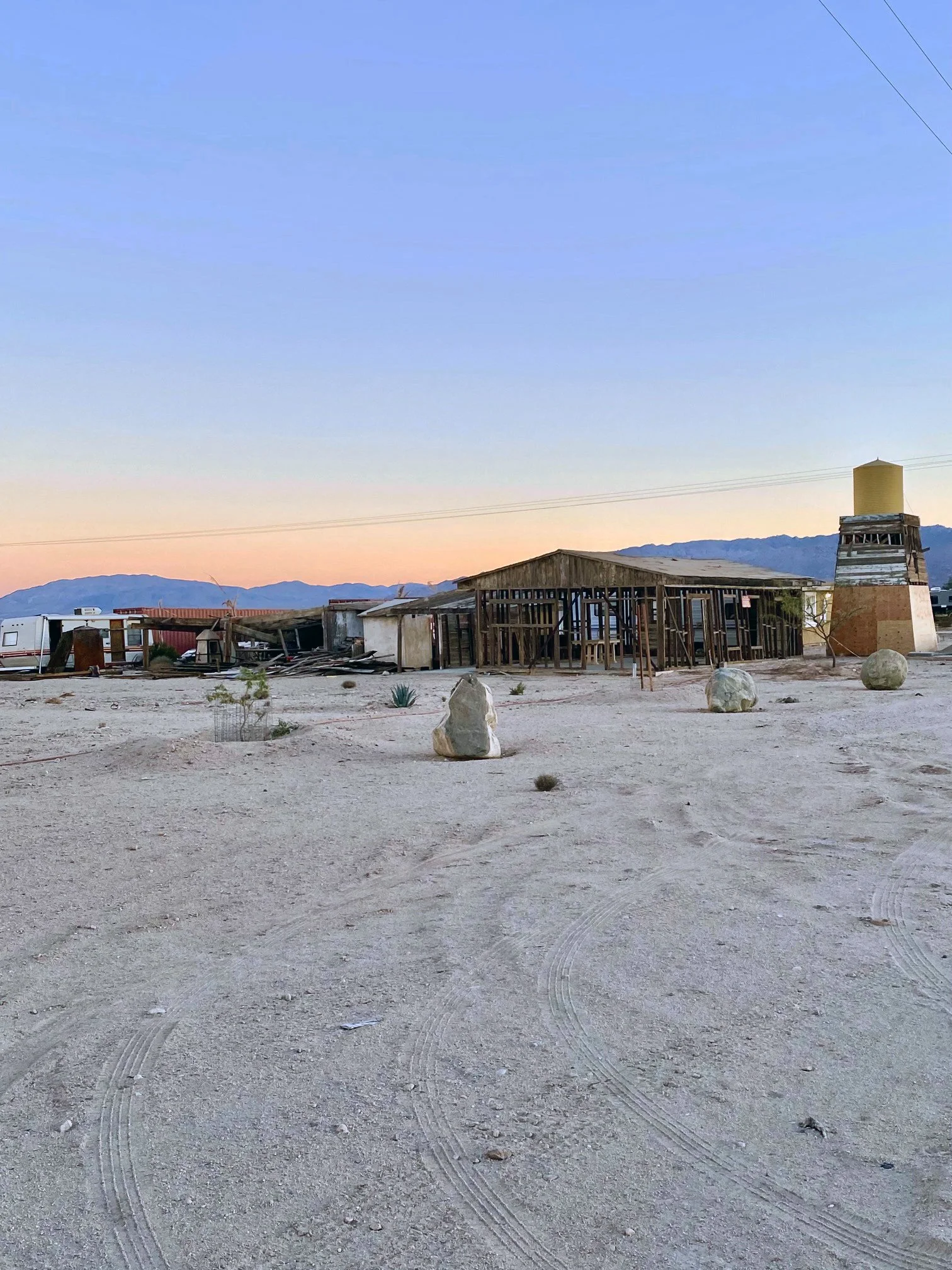 Desert landscape with abandoned wooden structures, large rocks, sparse vegetation, and distant mountains at sunset.