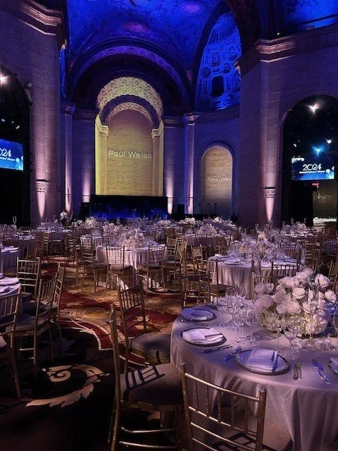 Elegant banquet hall with high arched ceilings illuminated in purple and gold, decorated with tables set for a formal event with white tablecloths, floral centerpieces, and chairs arranged around them.