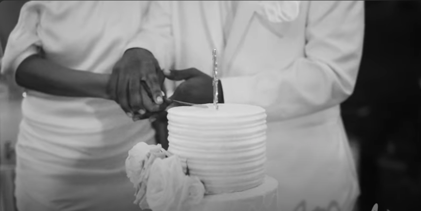 Two people cutting a birthday cake together, with a lit candle on top, in black and white.