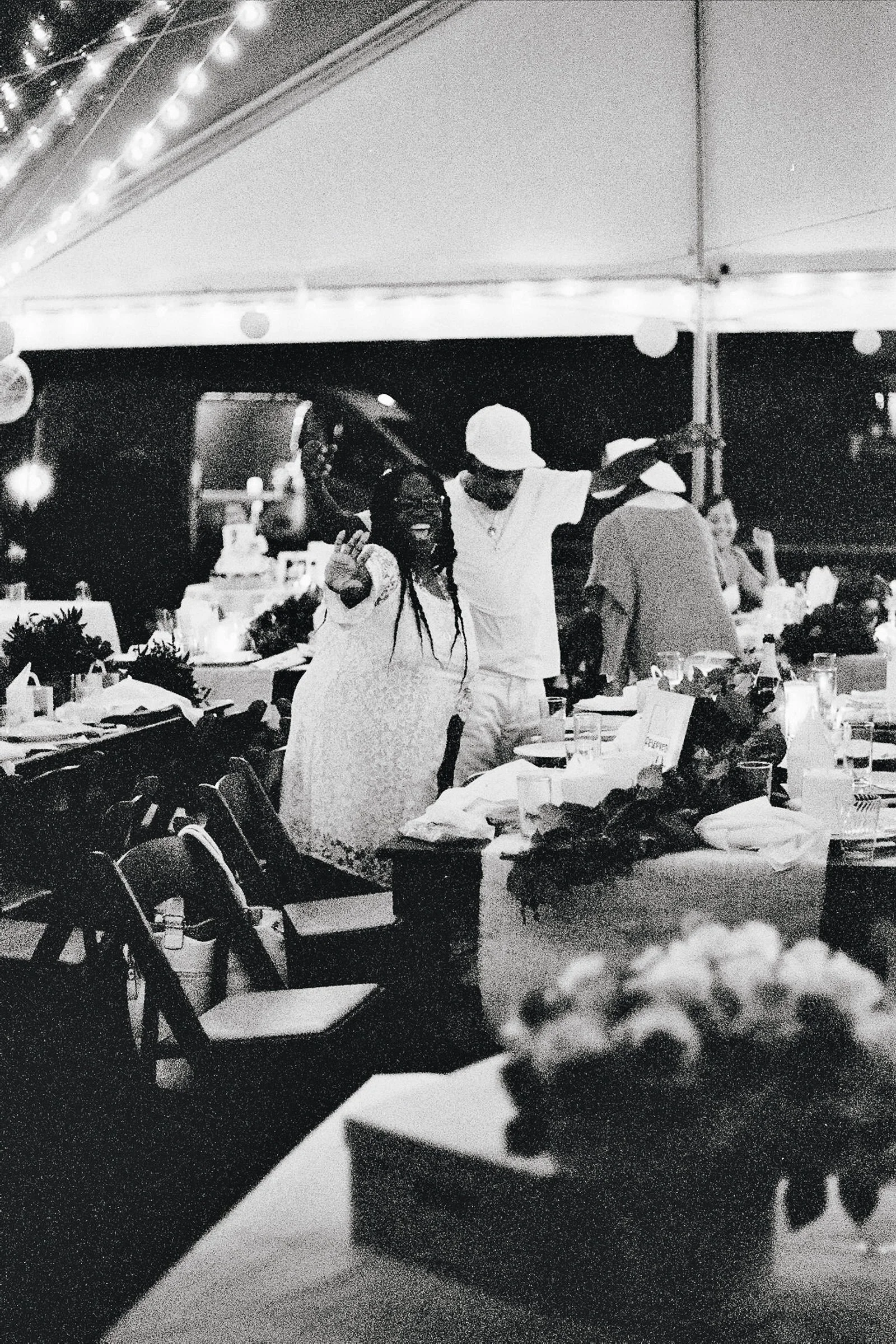 People celebrating at an outdoor nighttime party under a tent with string lights, tables with decorations, and a wedding cake in the background.