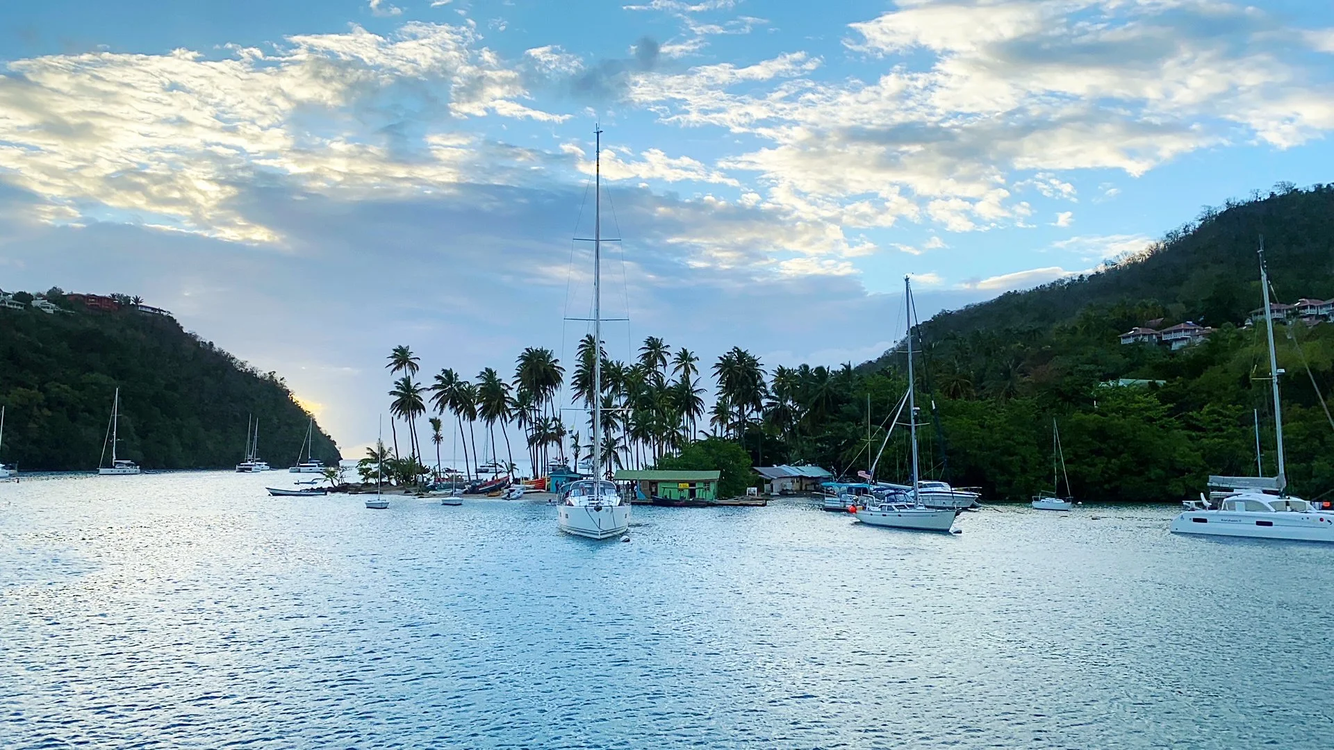 Sailboats anchored in a bay with lush green hills and a sky filled with clouds at sunset.