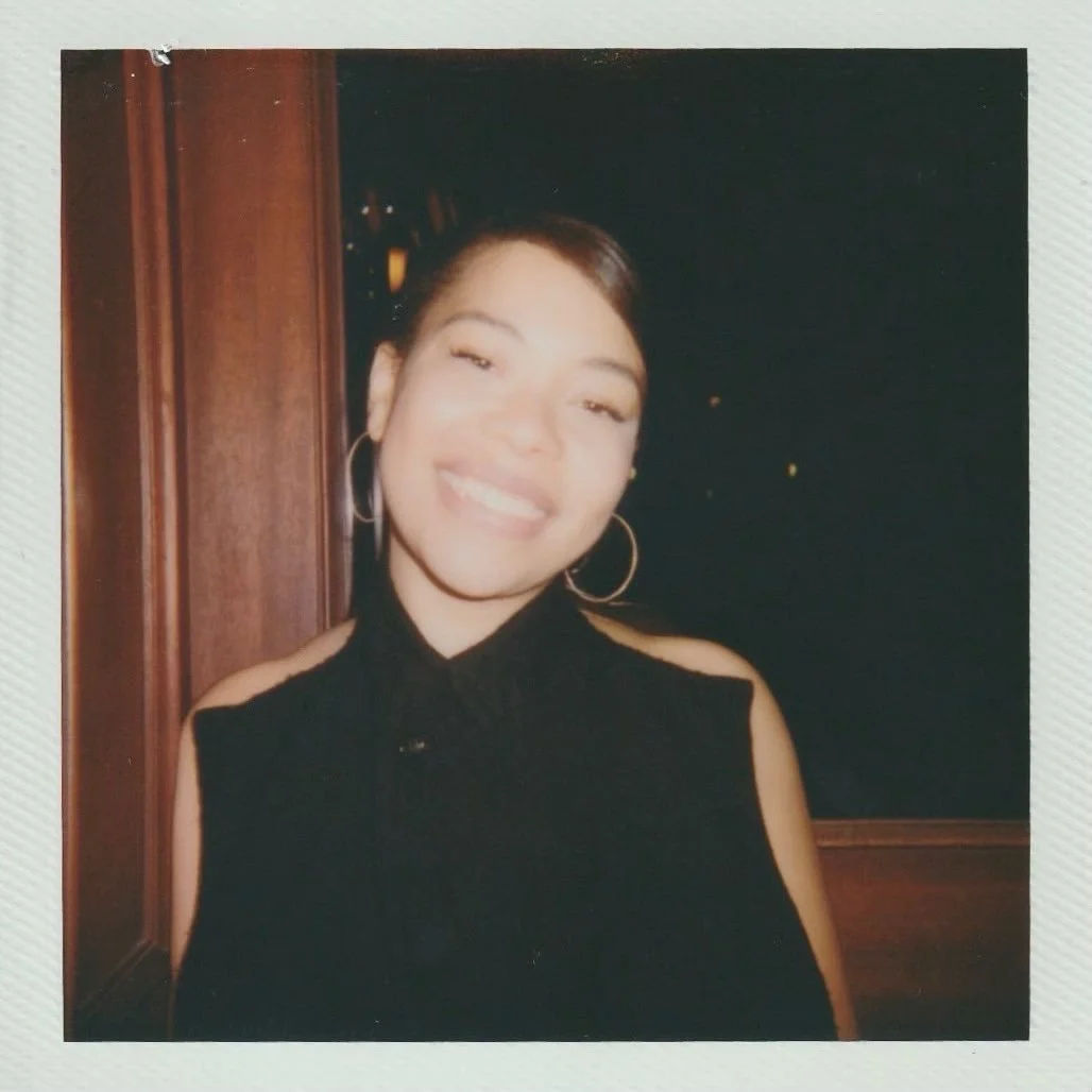Young woman smiling, wearing hoop earrings and a sleeveless black top, standing indoors near a wooden wall or partition.