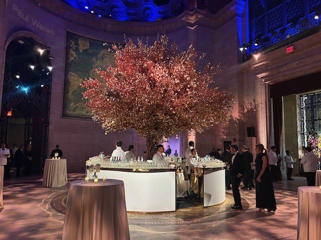 Elegant event with a large pink floral centerpiece, serving tables, and guests in formal attire.