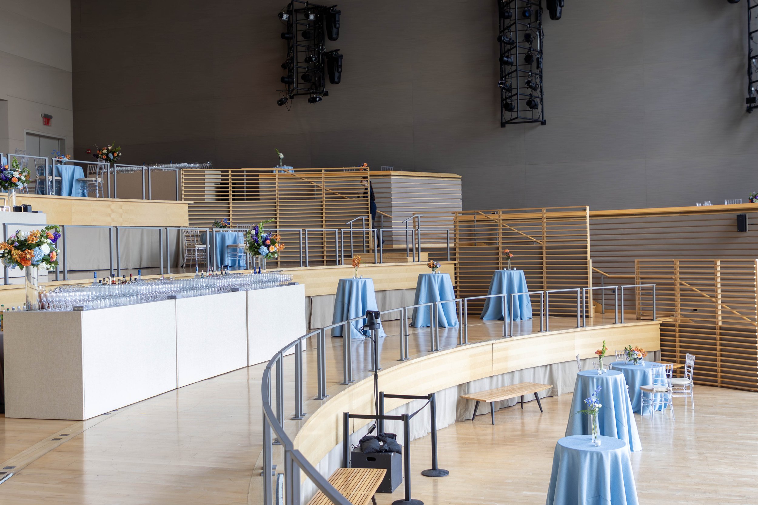 Empty event space with blue tablecloth-covered round tables, floral centerpieces, and a tiered platform with additional tables and floral arrangements, set up for a wedding or celebration.