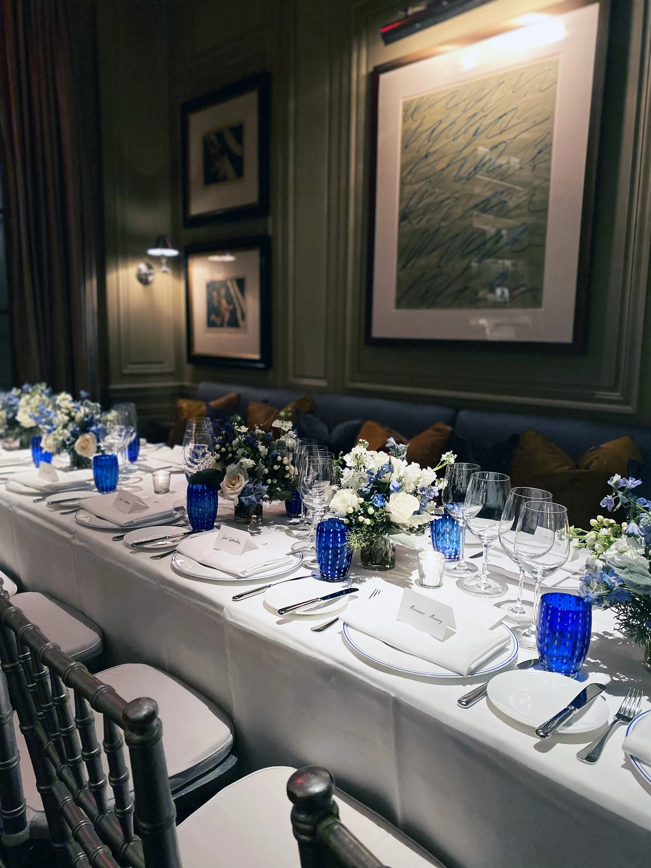 Elegant dining table set for a formal event with white tablecloth, floral centerpieces, blue glassware, and neatly arranged plates and utensils in a decorated room.