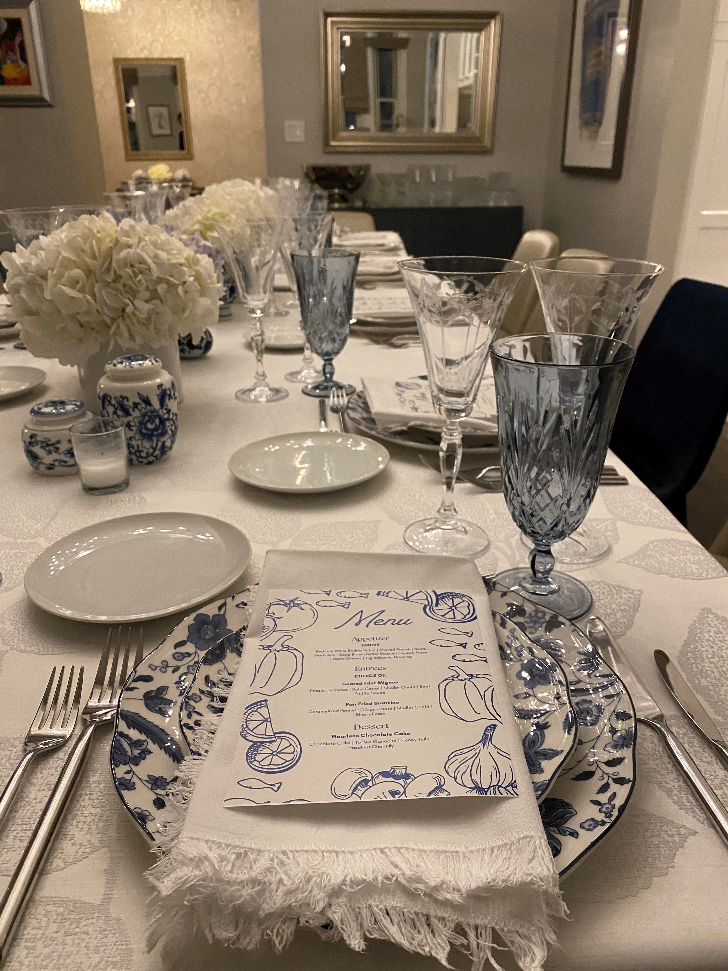 Set formal dining table with white and blue china, crystal glassware, a floral menu, white cloth napkin, and white floral centerpiece in a well-lit room with framed art on the walls.