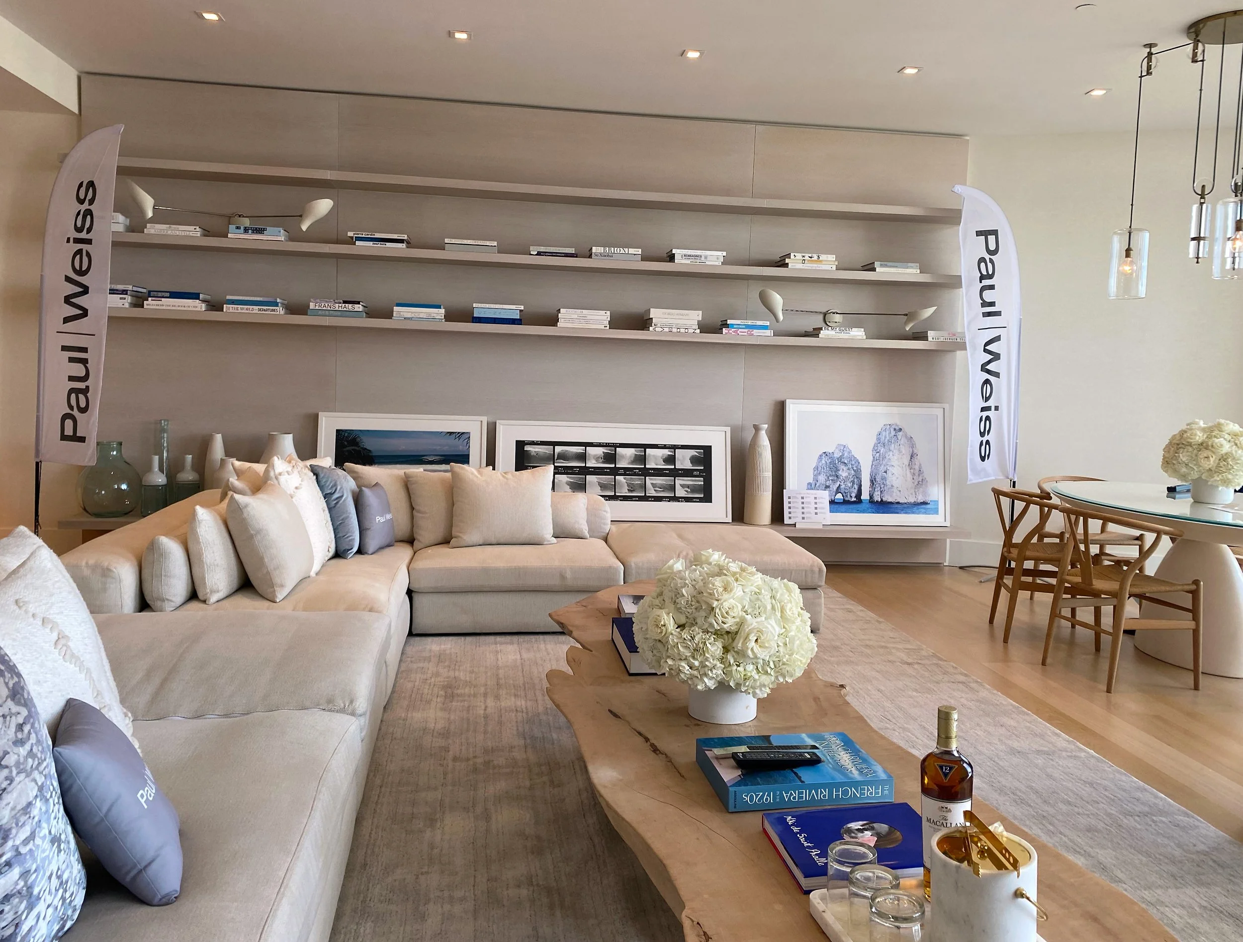 Living room with beige sectional sofa, white cushions, coffee table with flowers, books, bottle, and glasses, bookshelf on wall with books, two large framed photographs behind sofa, side table with vases, dining area with wooden chairs, floral centerpiece, and vertical banners with "Paul Weiss" on either side.