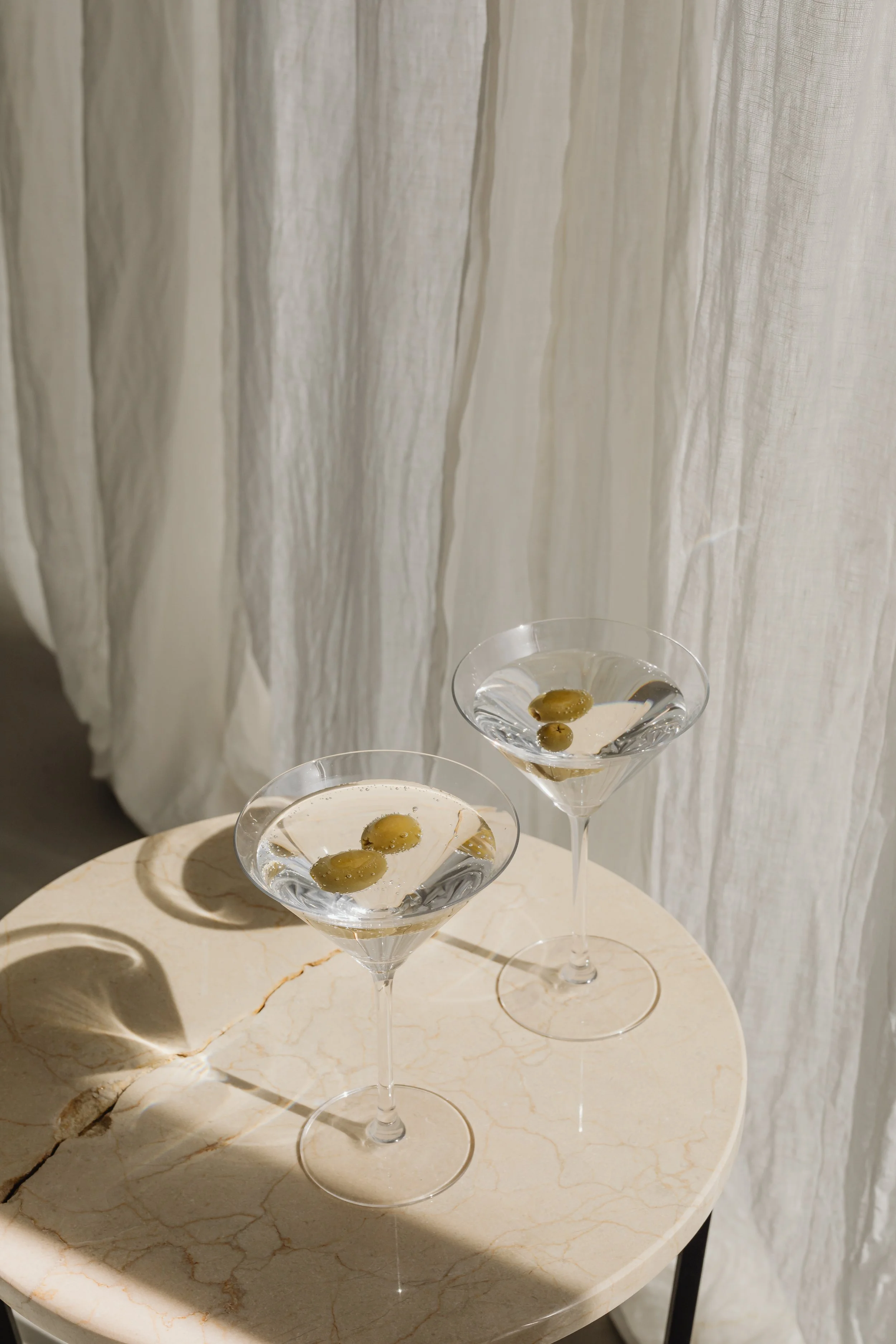 Two martini glasses filled with a clear liquid and green olives, placed on a round beige marble table near white curtains.