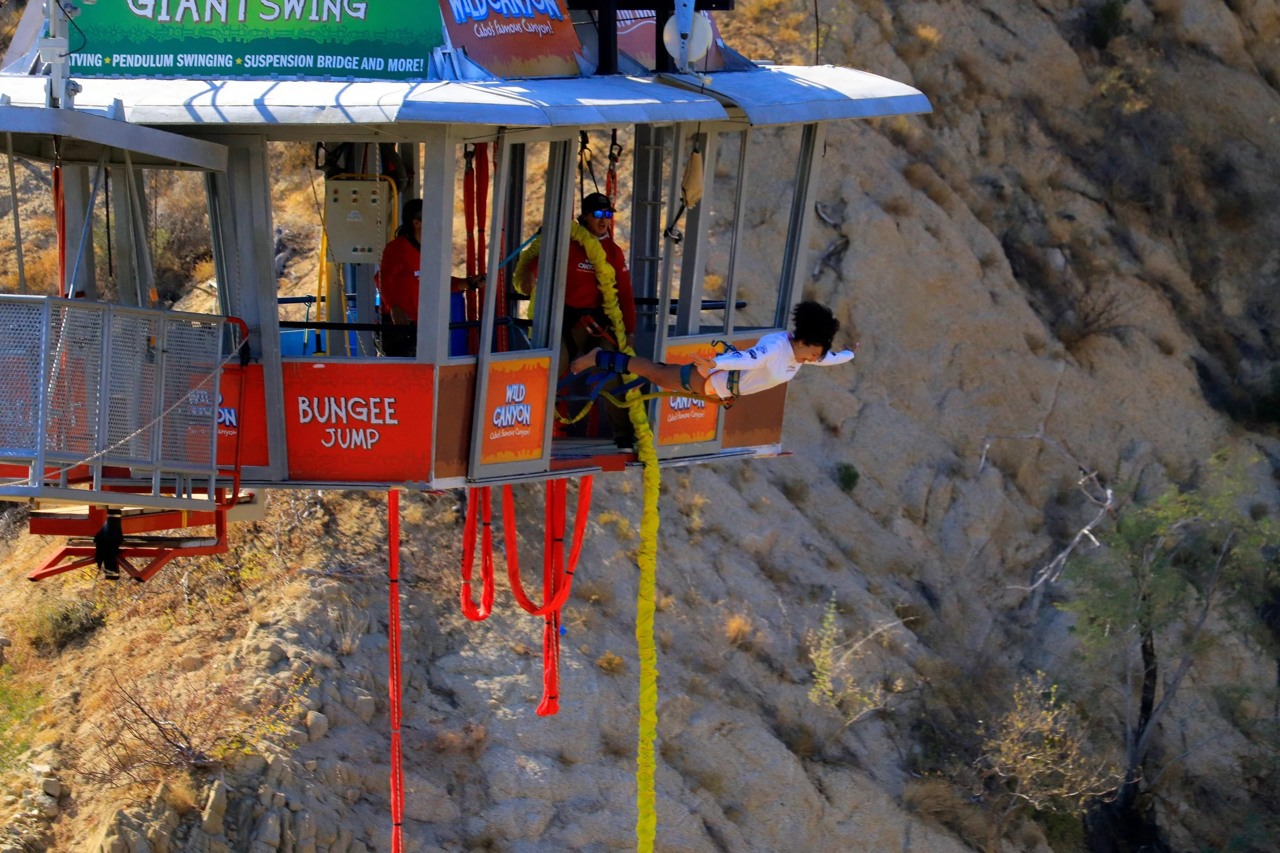 A person is jumping off a platform at Wild Canyon, a bungee jump attraction. The platform has a sign that says 'Bungee Jump' and is suspended over a rocky canyon landscape.