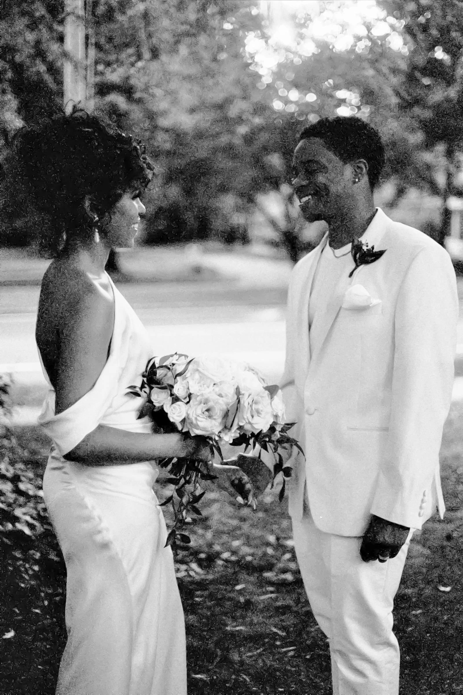 A black and white photo of a bride and groom standing outdoors, facing each other and smiling. The bride is holding a bouquet of flowers, and the groom is wearing a white suit with a black bow tie and a flower on his lapel. The background shows trees and a street.