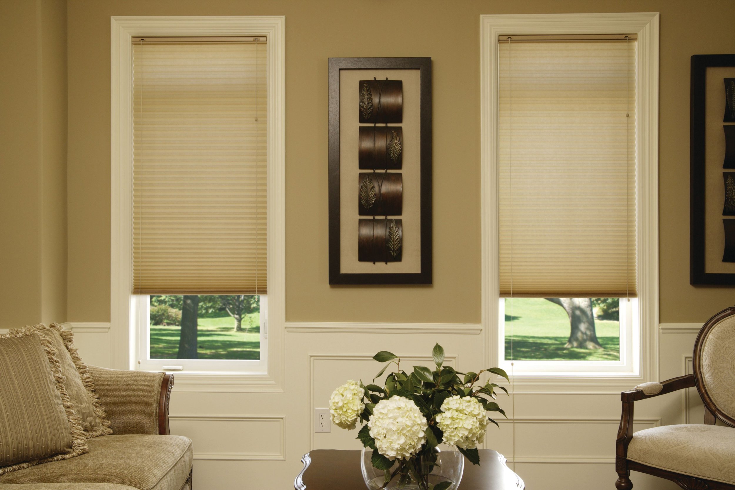 Living room with beige walls, two windows with closed blinds, a couch with pillows, a side chair, a coffee table with a bouquet of white flowers, and framed wall art.
