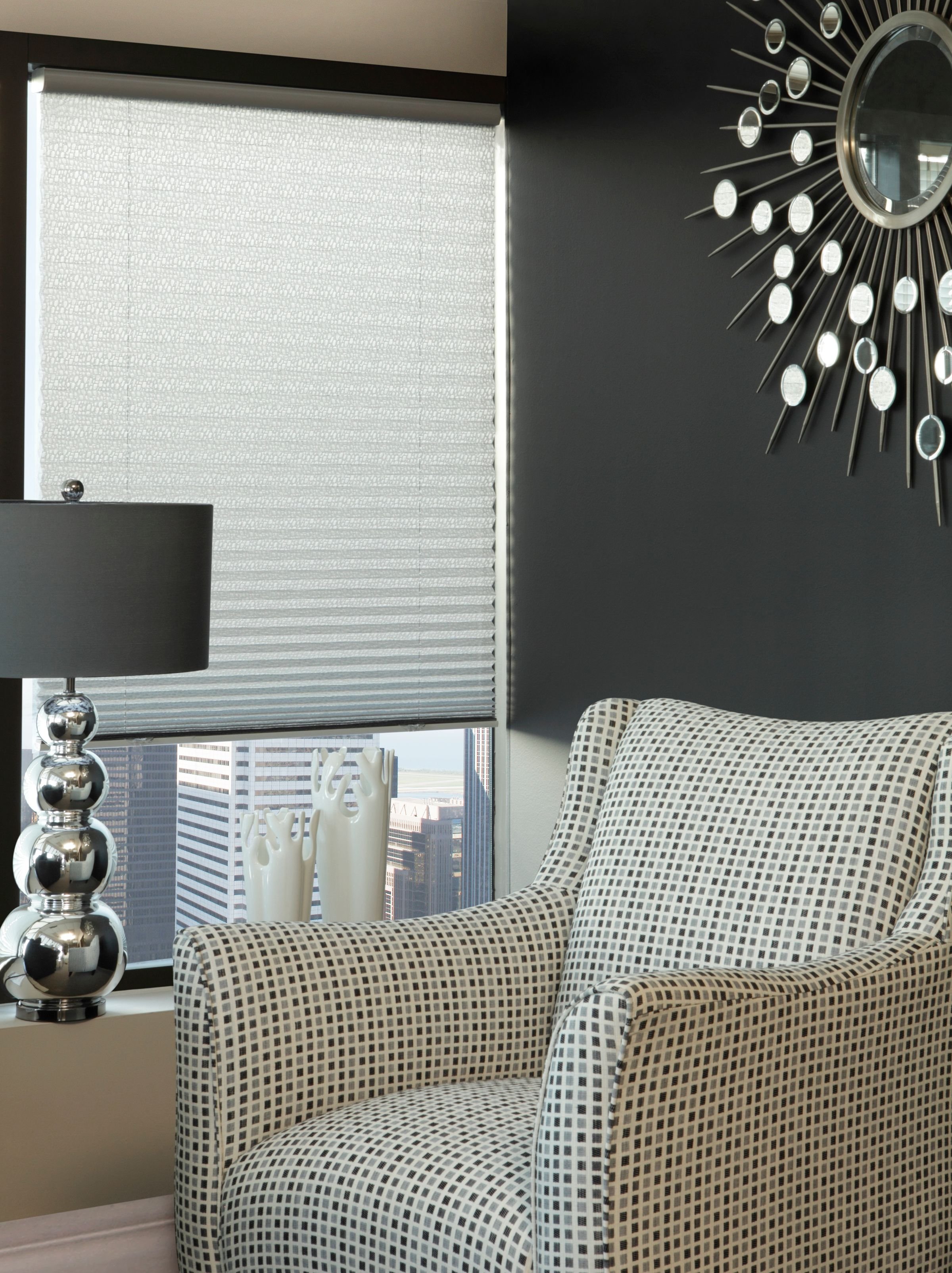 A stylish office corner with a checkered armchair, a lamp with a shiny metallic base, a decorative mirror with a sunburst frame, and a window with blinds showing a cityscape view.