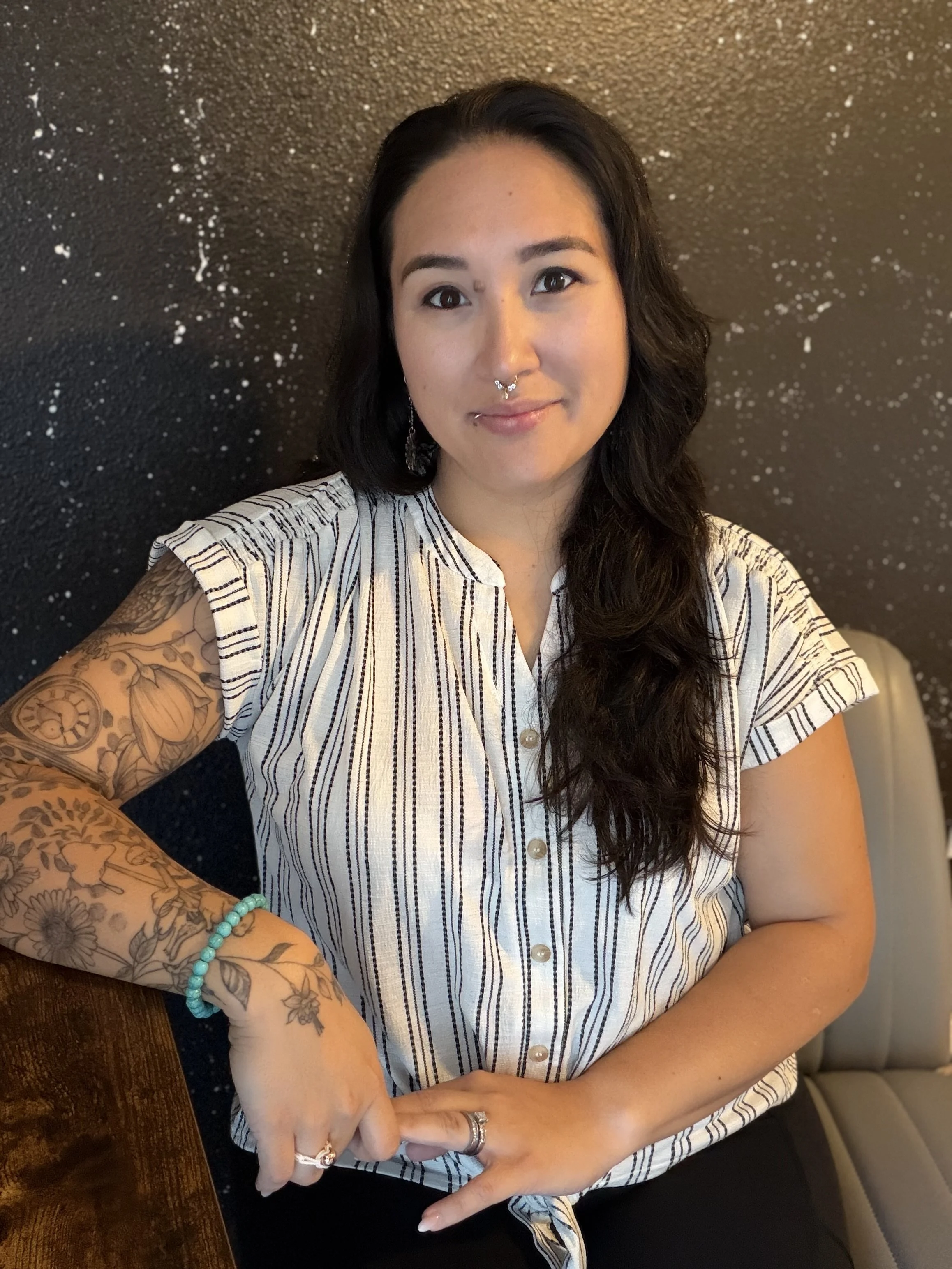 A woman with long dark hair and tattoos on her right arm, wearing a striped button-up shirt, gold hoop earrings, a septum piercing, and a nose piercing, sitting at a wooden table against a black wall with white speckles, pointing to her tattooed arm.
