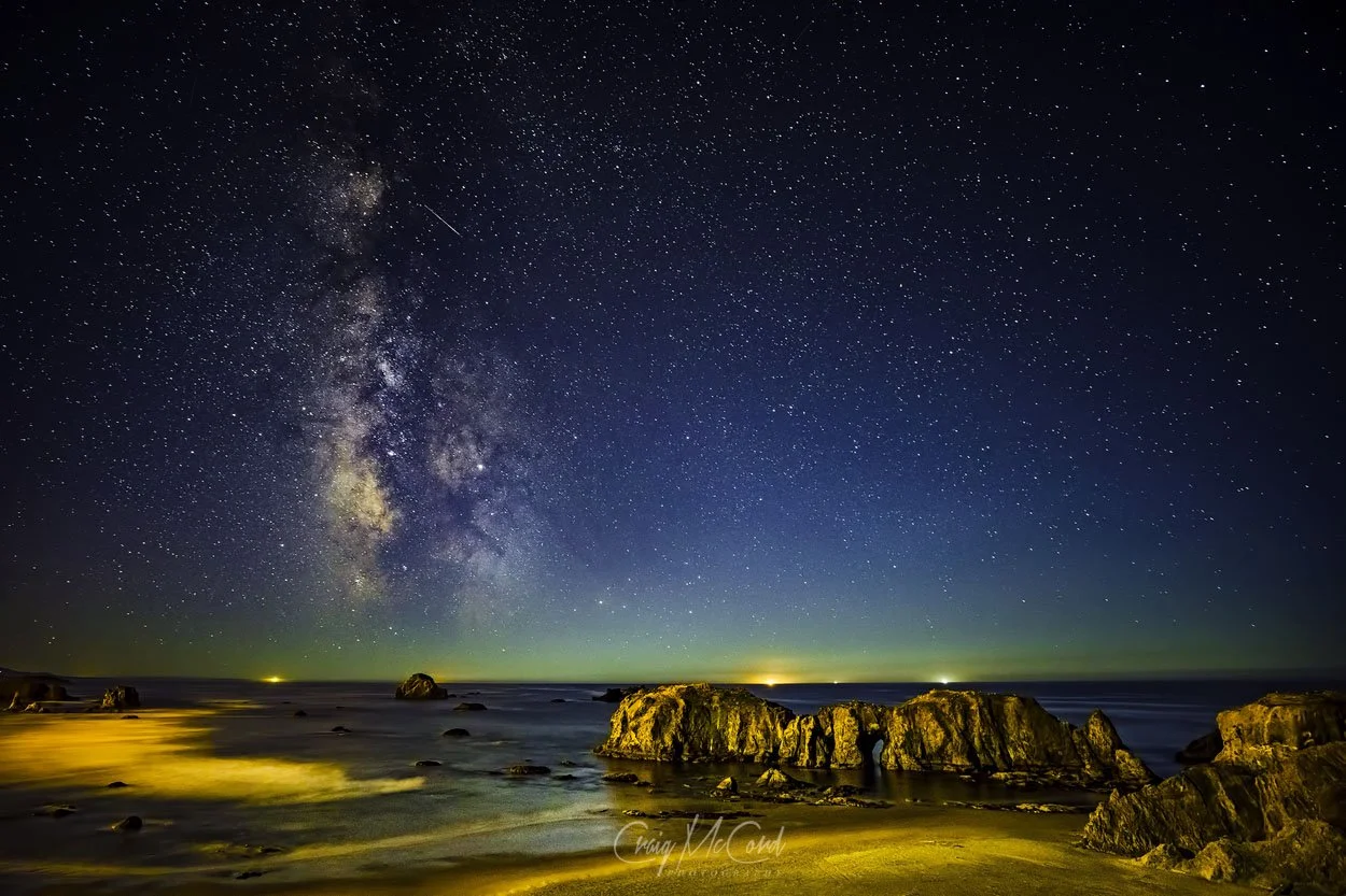 Night sky filled with stars and the Milky Way over a rocky beach with illuminated rocks and gentle waves at the shoreline.