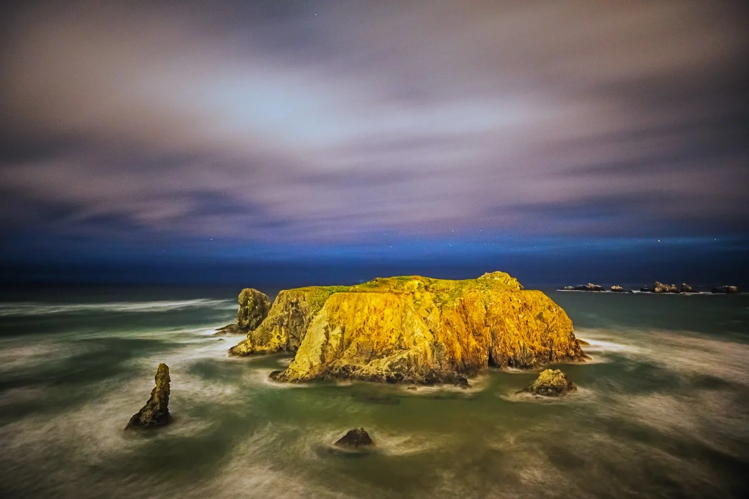 Nighttime coastal scene featuring a large moss-covered rock formation surrounded by smaller rocks in the ocean, with a cloudy sky and starry night sky in the background.