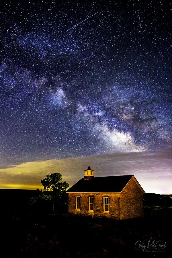 A starry night sky with the Milky Way galaxy and shooting stars above a small brick church with a pitched roof, surrounded by trees and dark landscape.