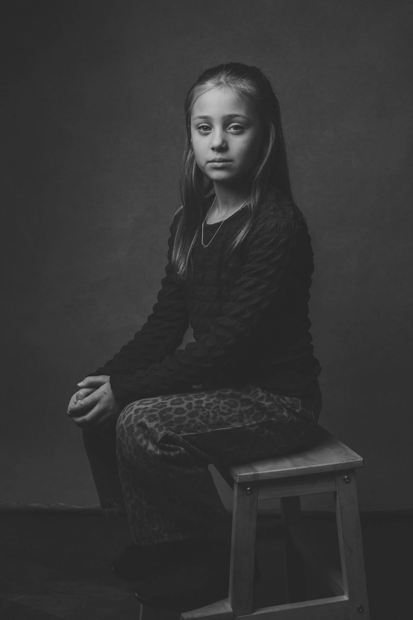 Black and white portrait of a young girl sitting on a stool, wearing a dark long-sleeve top and leopard print pants, with long hair and a neutral expression, against a dark background.