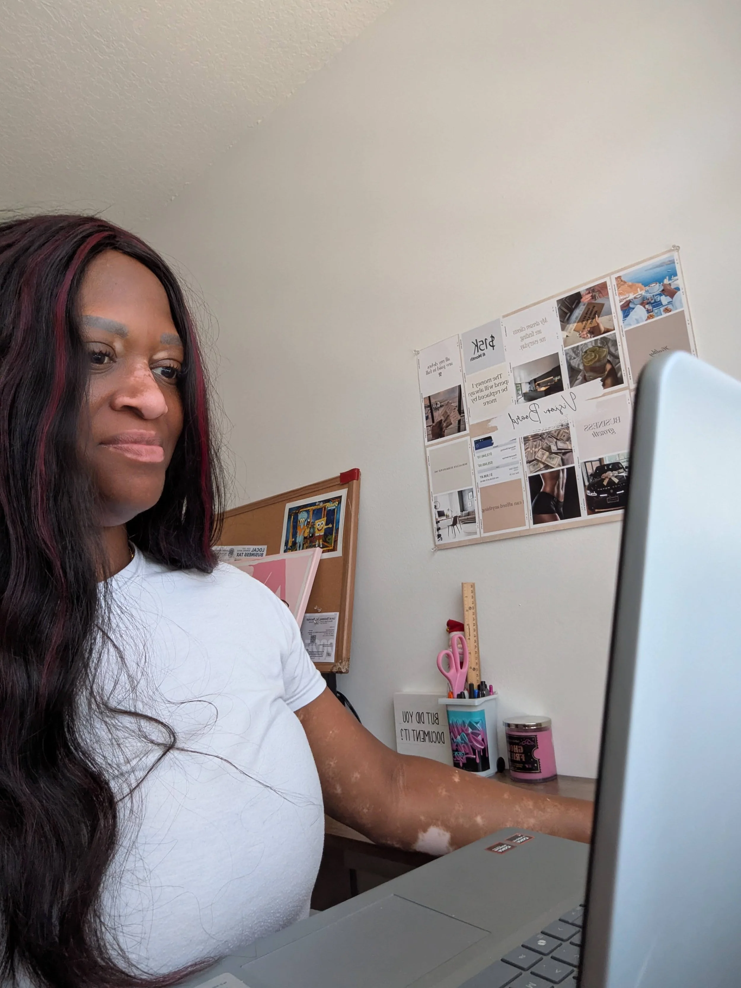a woman sitting at a computer with a vision board in the background