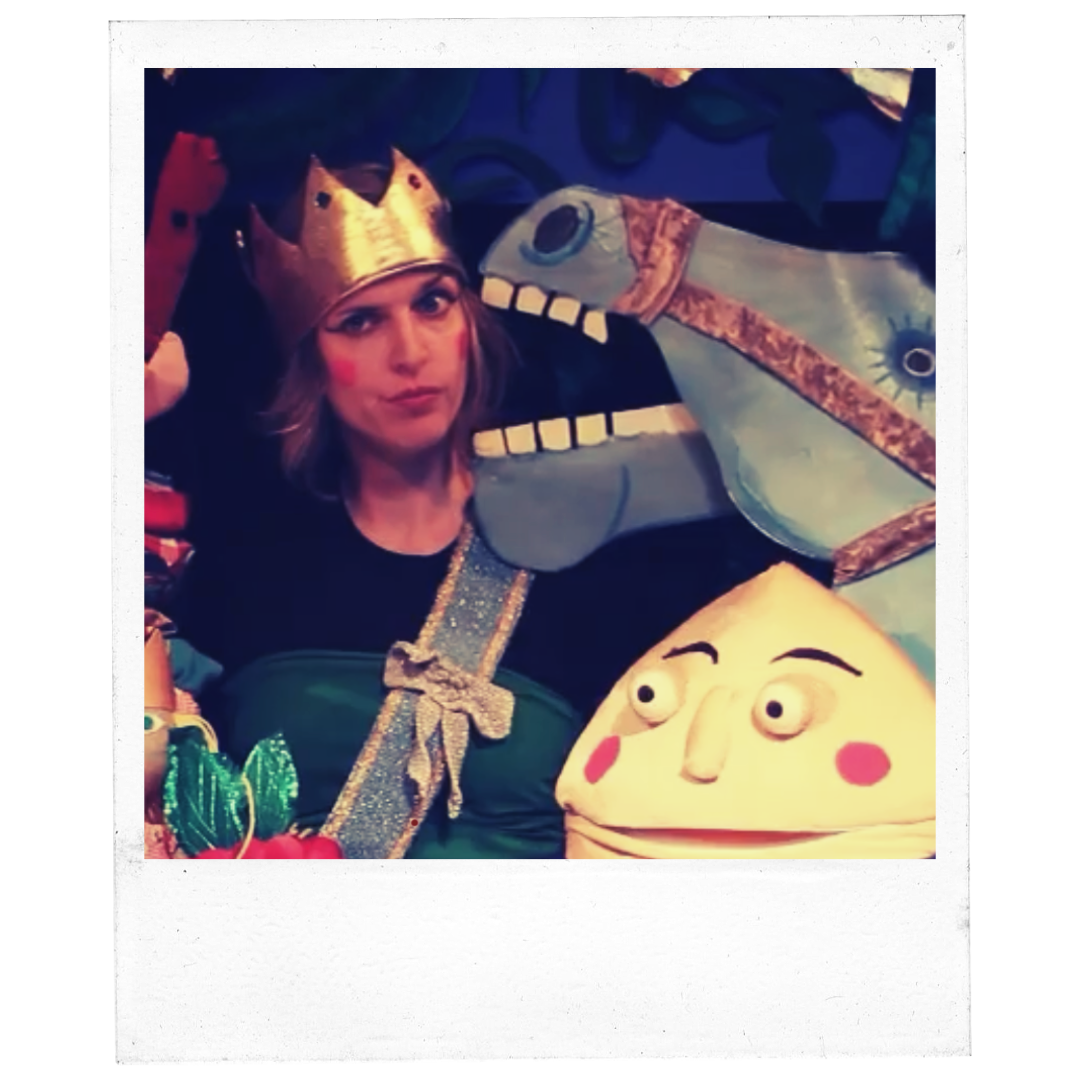 Person wearing a crown surrounded by colorful puppets, including a crocodile and a face-like character, in a playful setting.