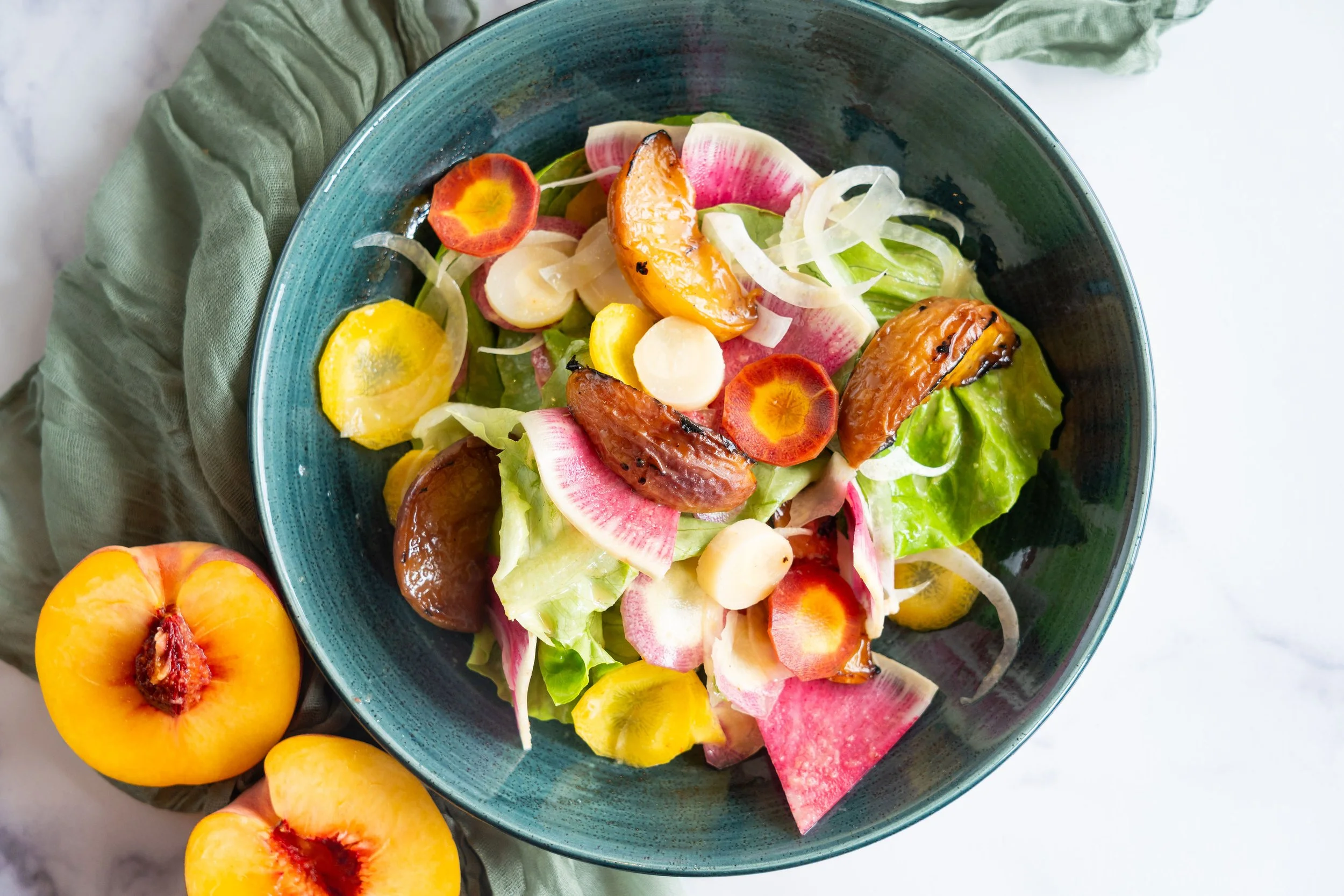 A green bowl of salad with grilled peaches, colorful radish slices, carrots, and greens, placed on a green cloth with fresh peaches beside it.