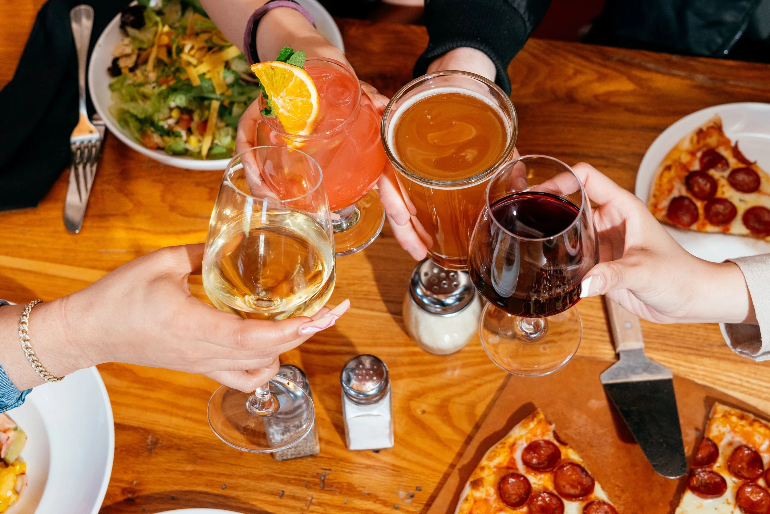 Four hands clinking glasses of wine, beer, and a cocktail over a wooden table with plates of salad and pepperoni pizza.