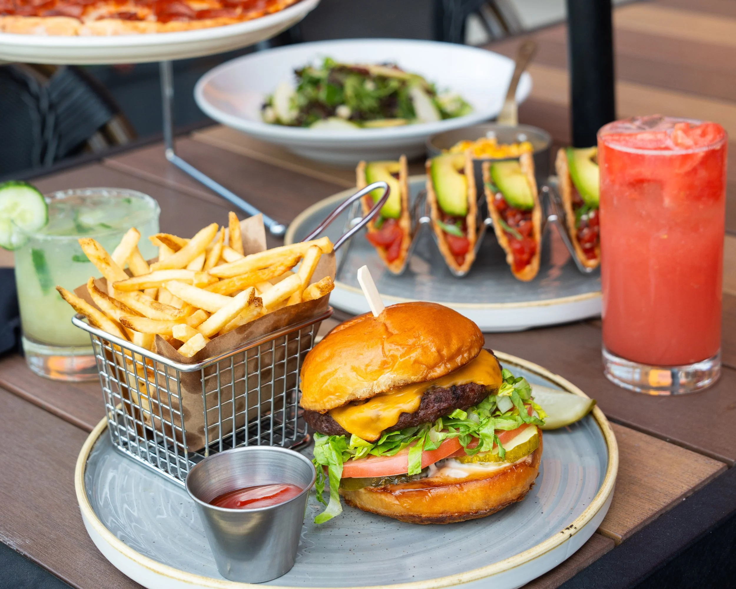 A plate with a cheeseburger topped with lettuce and tomato, served with fries and ketchup, alongside tacos, salad, pizza, and colorful drinks on a wooden table.