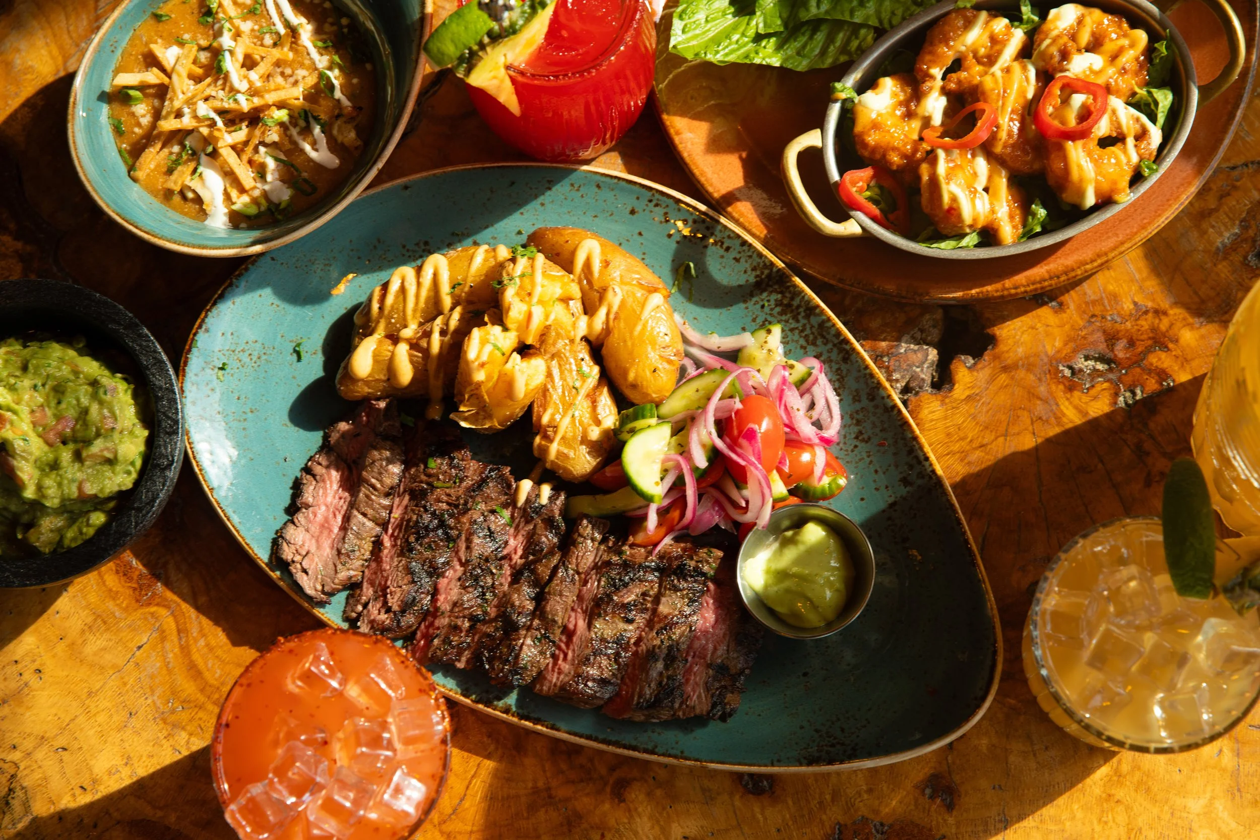 Plate of skirt steak with mushrooms and salad, surrounded by colorful cocktails, tortilla chips, and a bowl of soup on a wooden table.