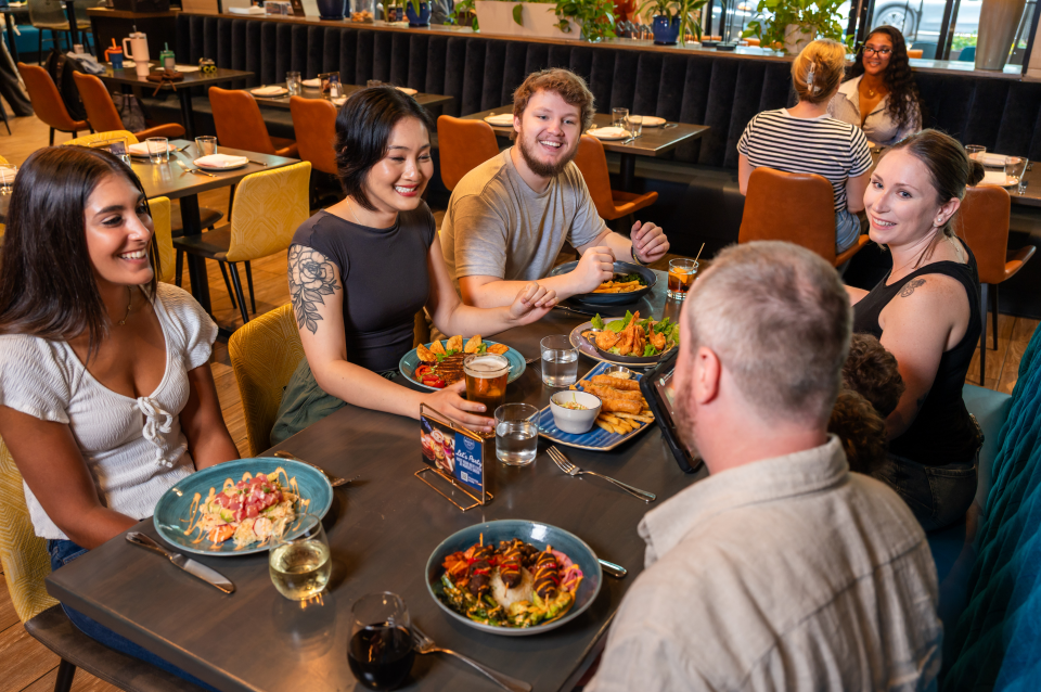 Group dining at a restaurant table with plates of colorful dishes, drinks, and appetizers in a modern setting.