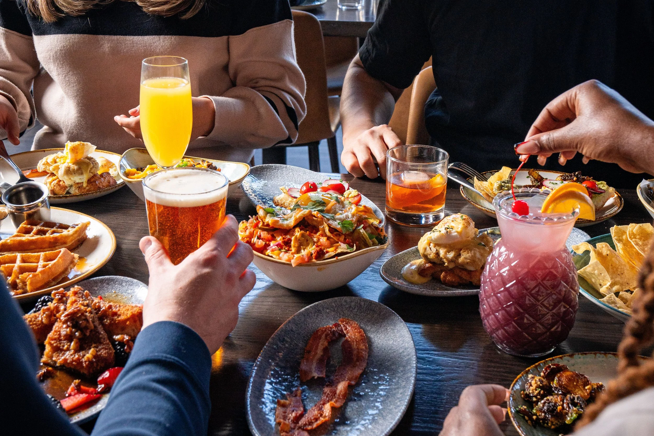 A group of people enjoying brunch with plates of waffles, fried chicken, bacon, and colorful cocktails on a dark wooden table.