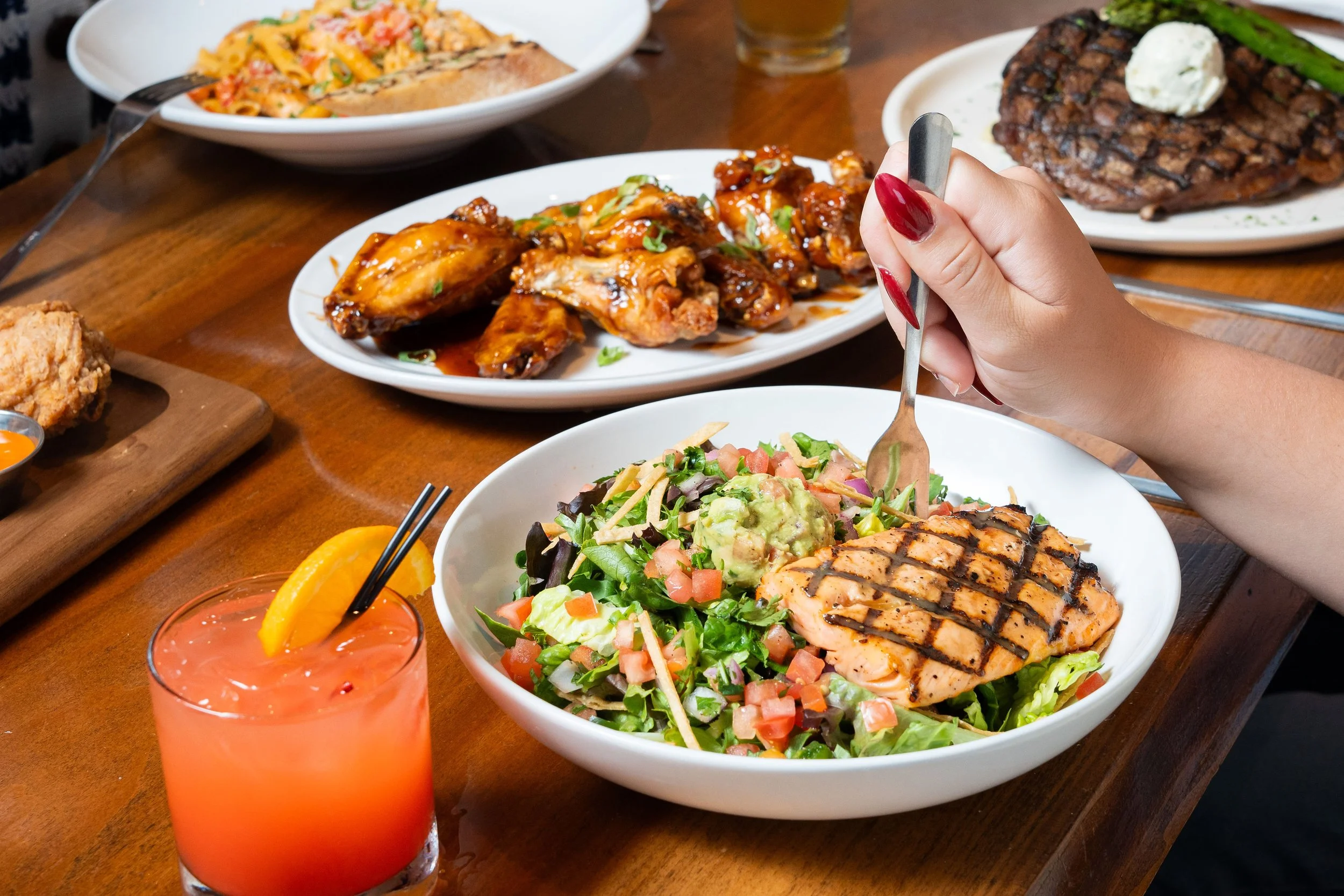 A hand holding a fork over a bowl of salad topped with grilled salmon and guacamole, with plates of wings, steak, and pasta, plus a bright orange cocktail on a wooden table.